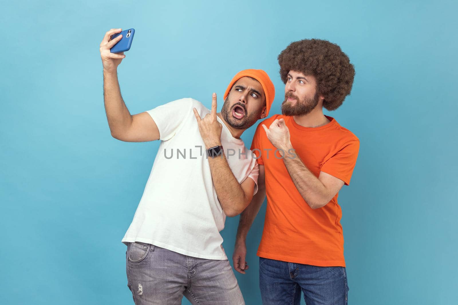 Portrait of funny cool two young adult hipster men blogger broadcasting livestream or taking selfie, showing rock and roll and shaka gestures. Indoor studio shot isolated on blue background.