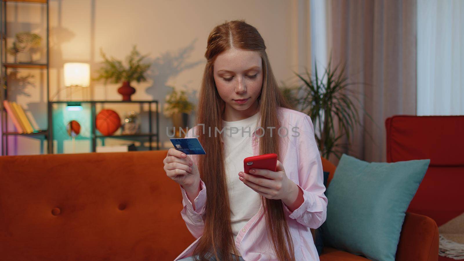 Happy shopaholic consumer young redhead teen child girl sitting at home making online order food delivery, payment with credit plastic bank card. Discount sale on internet. Ginger female children kid