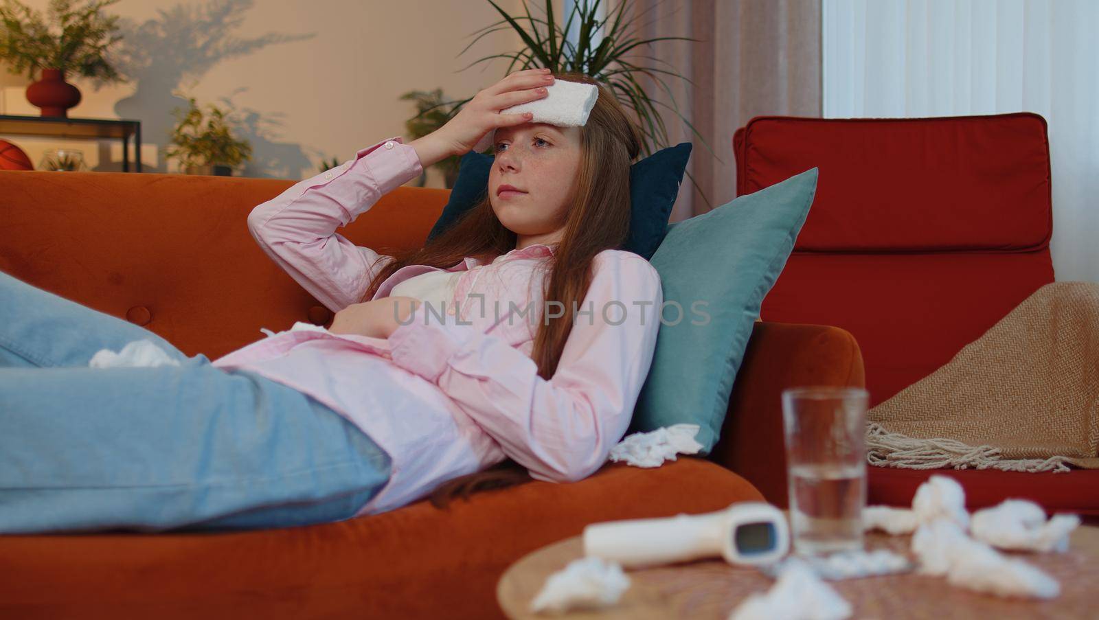 Young ill child kid suffering from cold or allergy lying on sofa in living room at home. Sick children girl with towel on head blows sneezes wipes snot into napkin. Coronavirus quarantine, pandemic