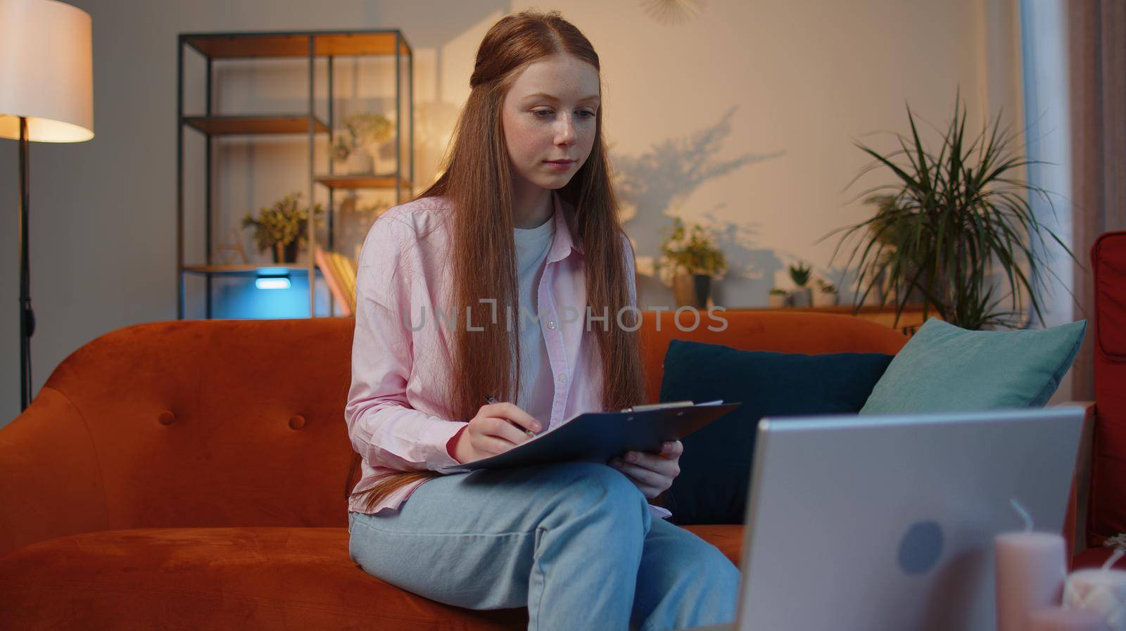 Happy smiling young child girl accounting payments and calculating monthly expenses bank bills, analysing paperwork graphs at home couch. Red hair kid freelancer working online remote job on laptop