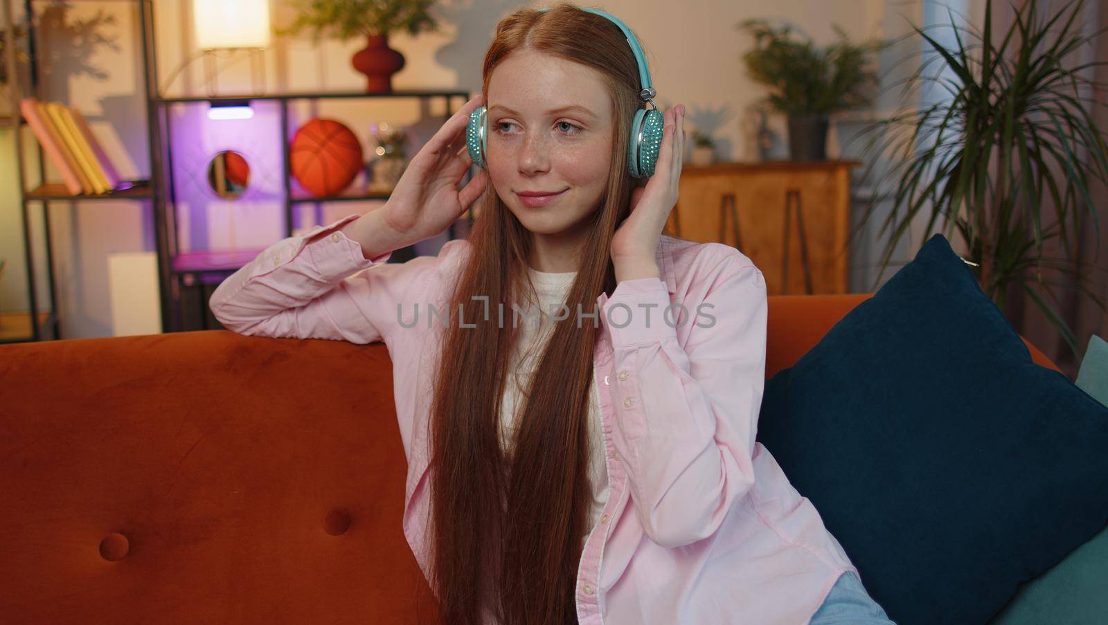 Happy relaxed overjoyed ginger kid in wireless headphones dancing on couch at home choosing listening favorite energetic disco rock n roll music in smartphone entertaining. People weekend activities