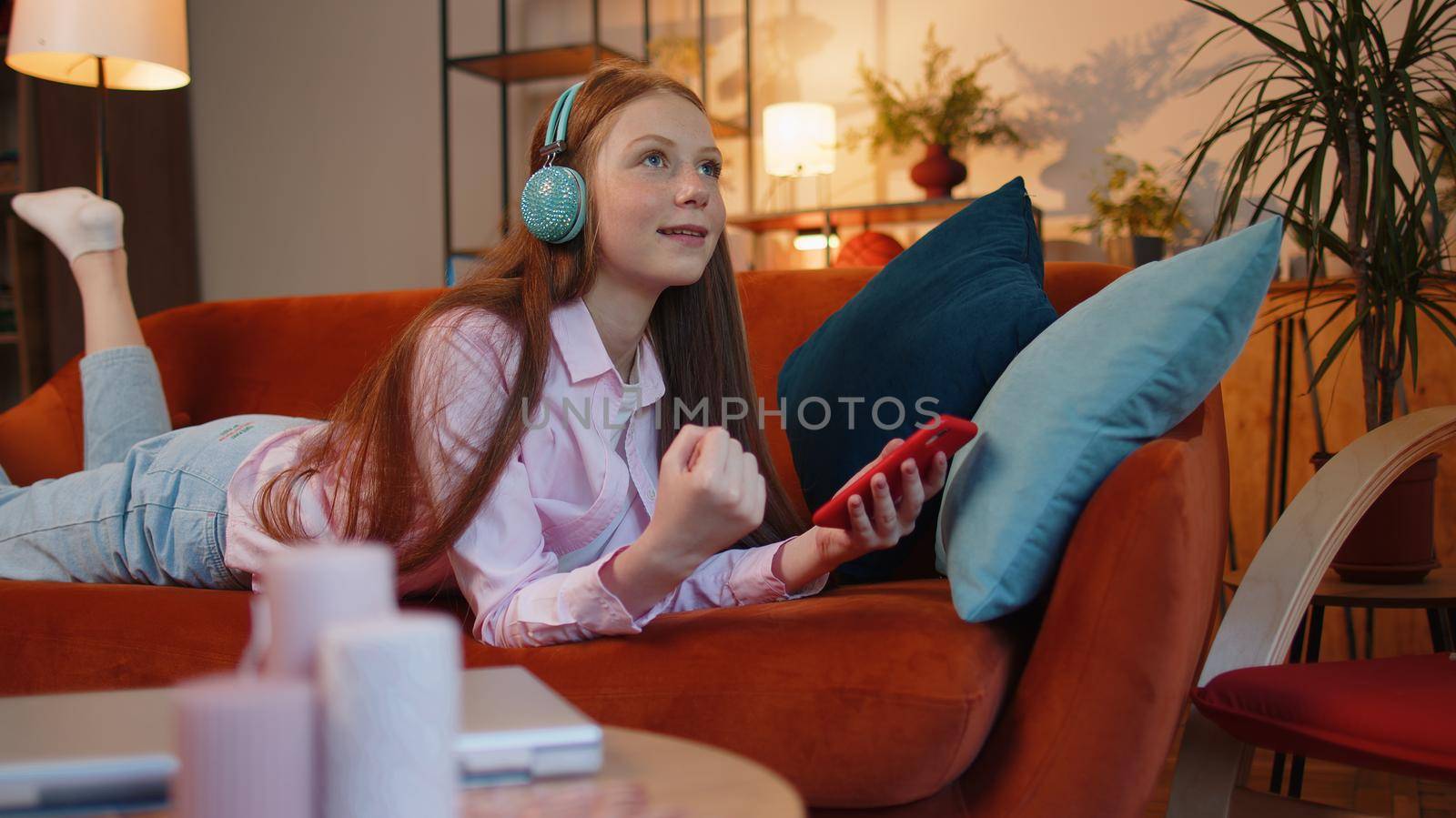 Happy excited joyful young redhead teenager child girl use mobile smartphone typing browsing say Wow yes found out great big win good news celebrate sitting at home couch. Ginger female children kid