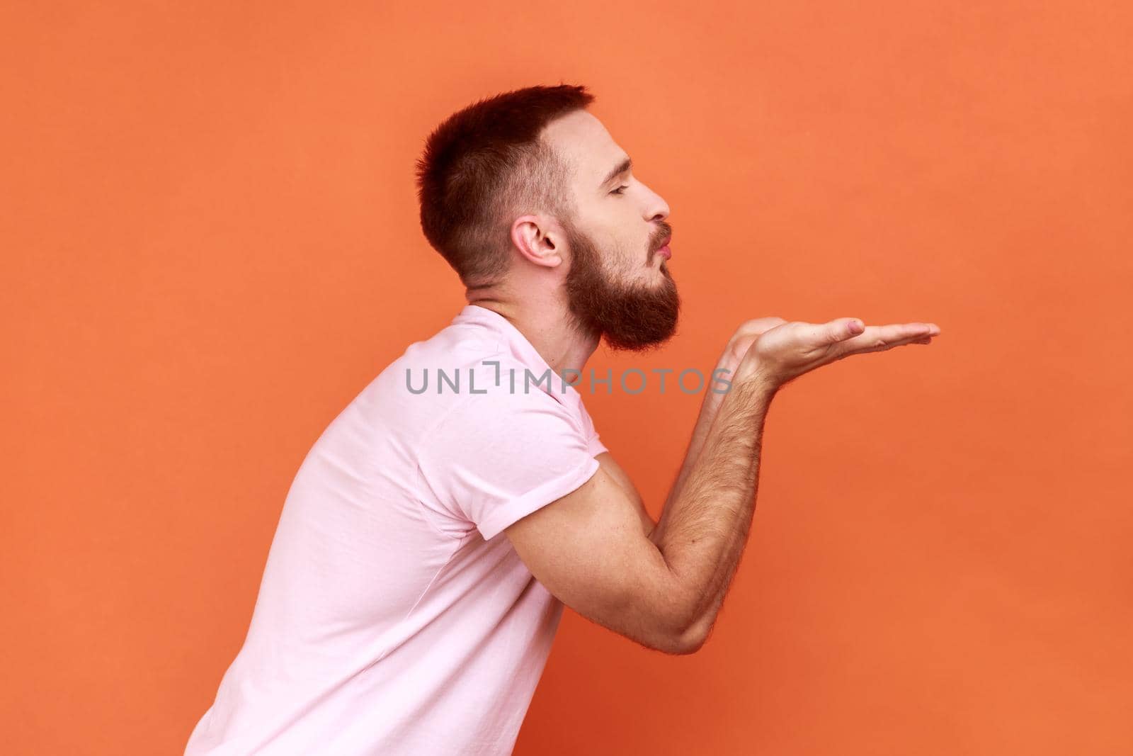 Side view portrait of handsome romantic young adult bearded man standing and looking ahead and sending air kiss, wearing pink T-shirt. Indoor studio shot isolated on orange background.