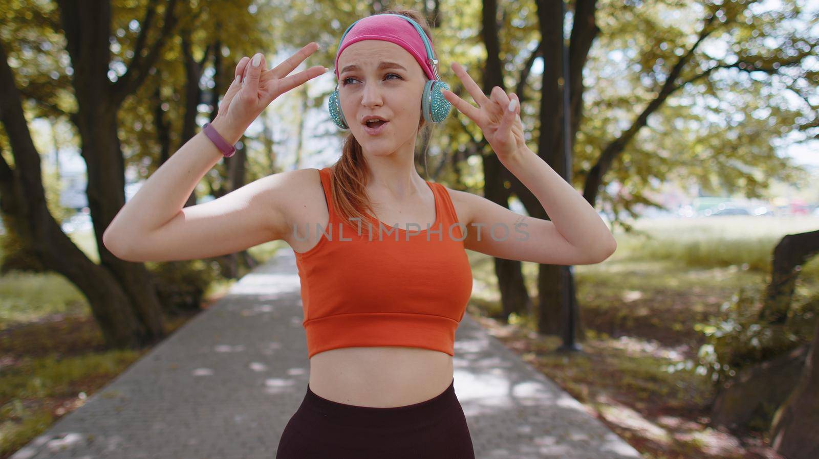Athletic fitness sport runner girl training exercise, listening music on headphones dancing to camera having fun. Workout cardio in park. Young woman jogger enjoying funny dance. Active sportswoman