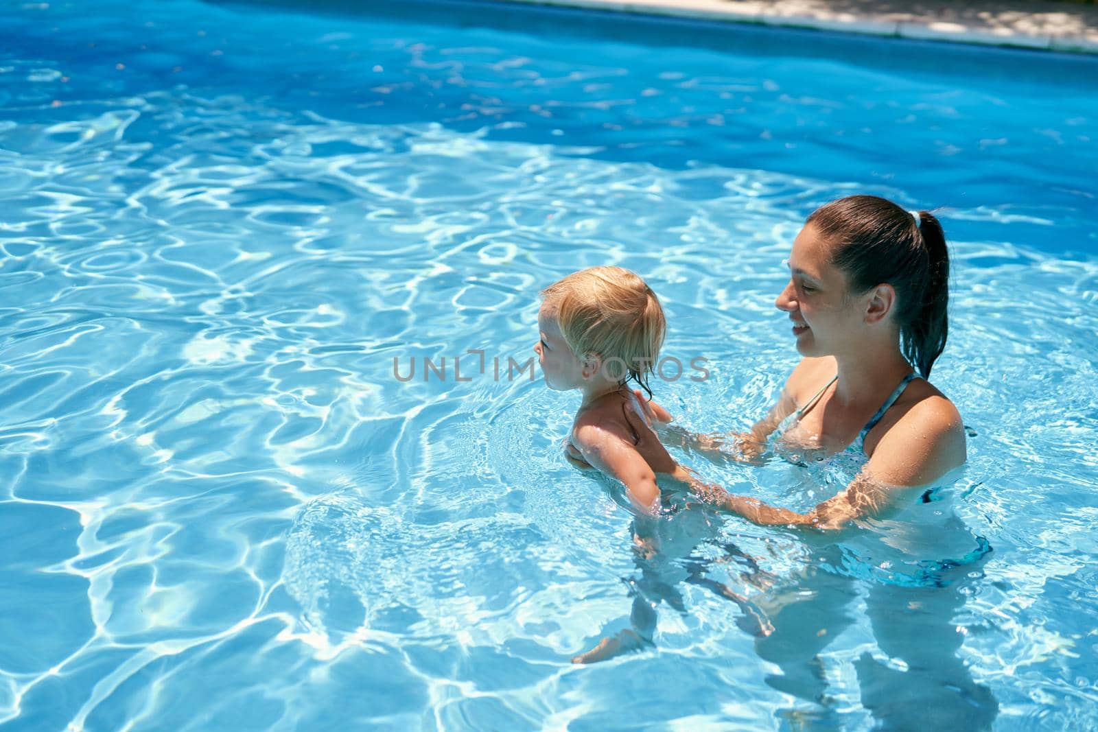 Mom teaches a small baby to swim in a pool with turquoise water by Nadtochiy