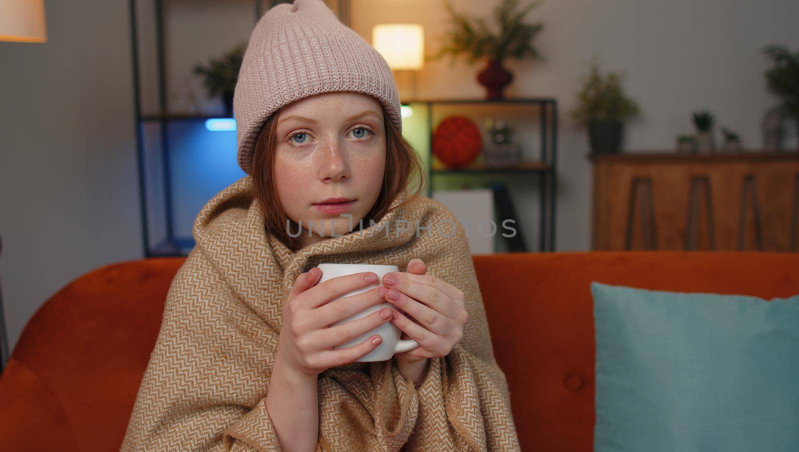 Sick child girl wear hat wrapped in plaid sit alone shivering from cold on sofa drinking hot tea in unheated apartment without heating due debt. Unhealthy children feeling discomfort try to warming up
