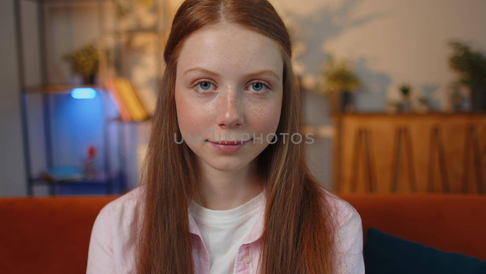 Close-up portrait of happy positive smiling young redhead child girl, looking at camera. Teen red-haired ginger freckles face kid indoor isolated at home in living room on couch. Female nature beauty