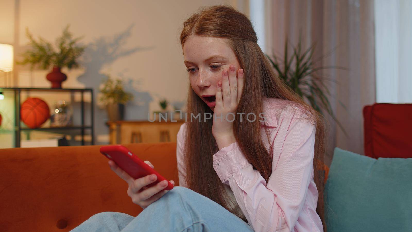 Oh my God, Wow. Happy excited teen young child girl use mobile smartphone typing browsing say yes found out great big jackpot lottery win, good news, celebrate at home couch. Ginger children kid alone