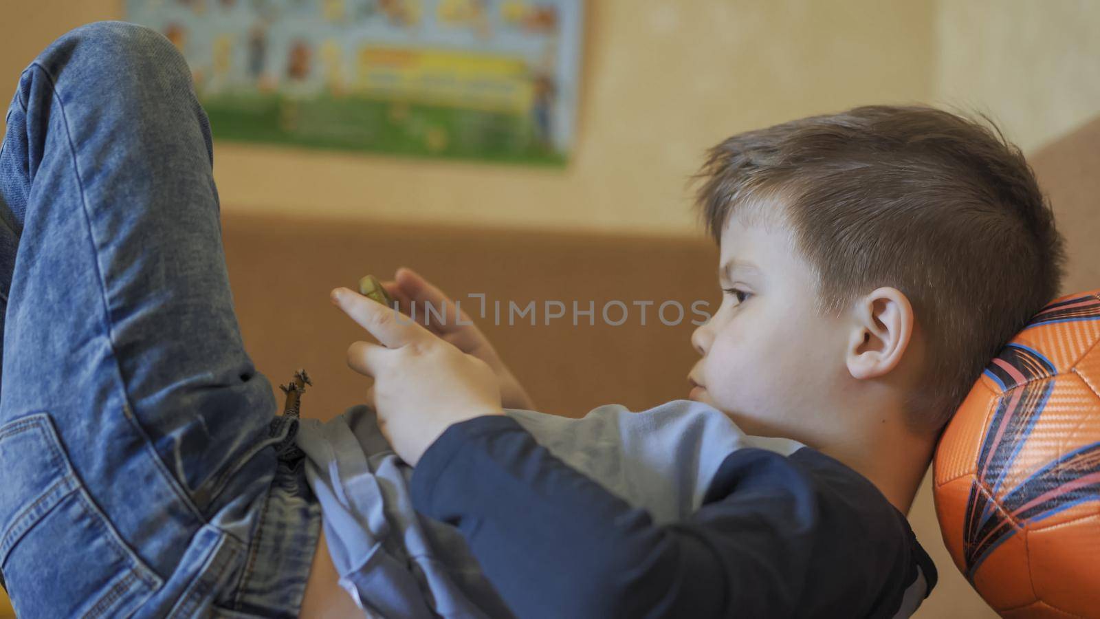 Boy Plays On Mobile Phone Lying On Sofa, Kid Uses Orange Ball Instead Of Pillow, The Boy Is Forced To Stay At Home In Isolated Due To Coronavirus Epidemic, Profile View