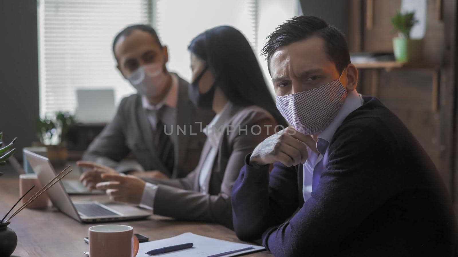 Business People In Protective Masks At Meeting In Office Despite Quarantine, Focus On Serious Young Man Communicates With Colleagues Sitting At Table In Board Room