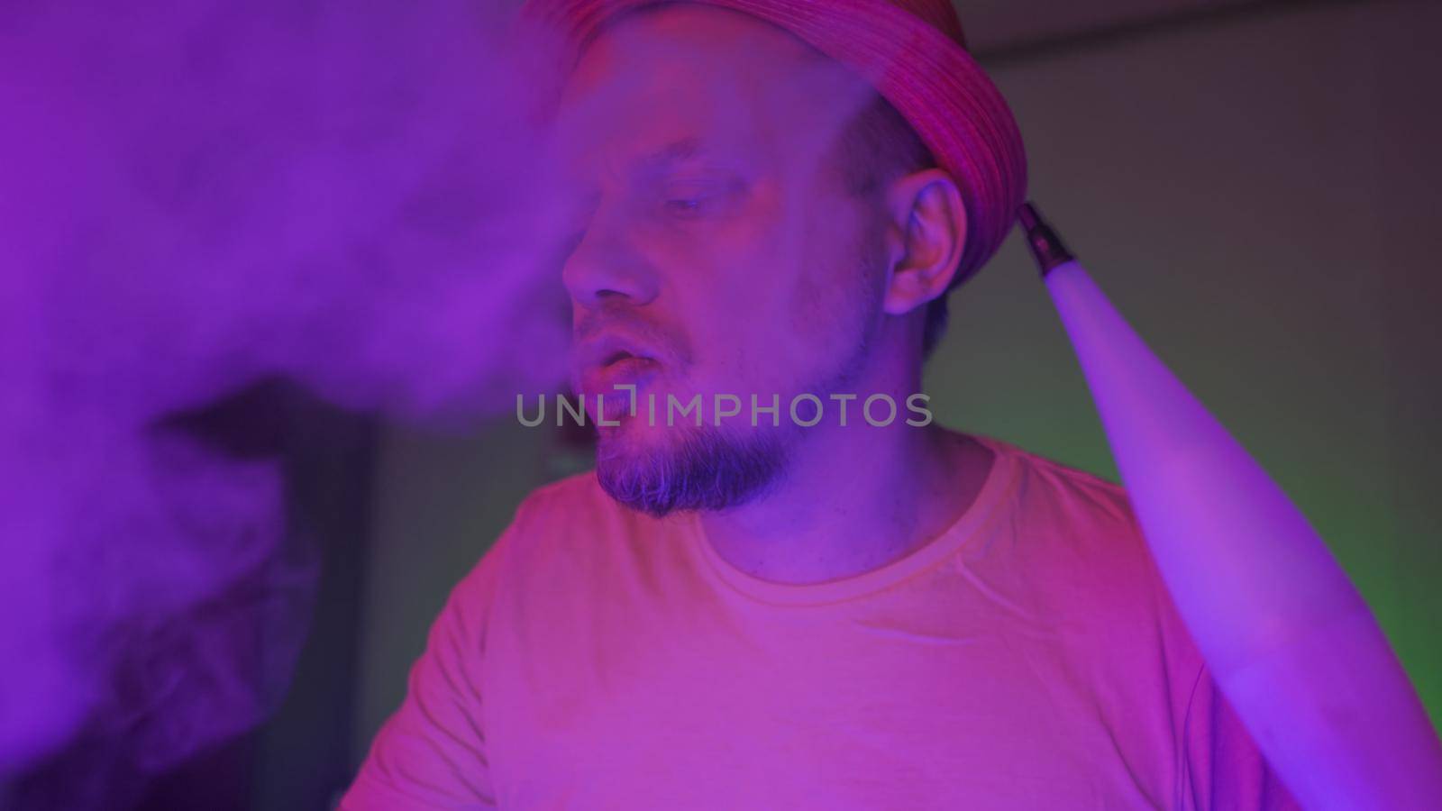 Smoker Of Hookah Exhales Smoke In Neon Lighting, Creative Man In Hat Smoking Hookah Sitting In Foggy Dark Room With Fluorescent Illumination, Quarantine Concept