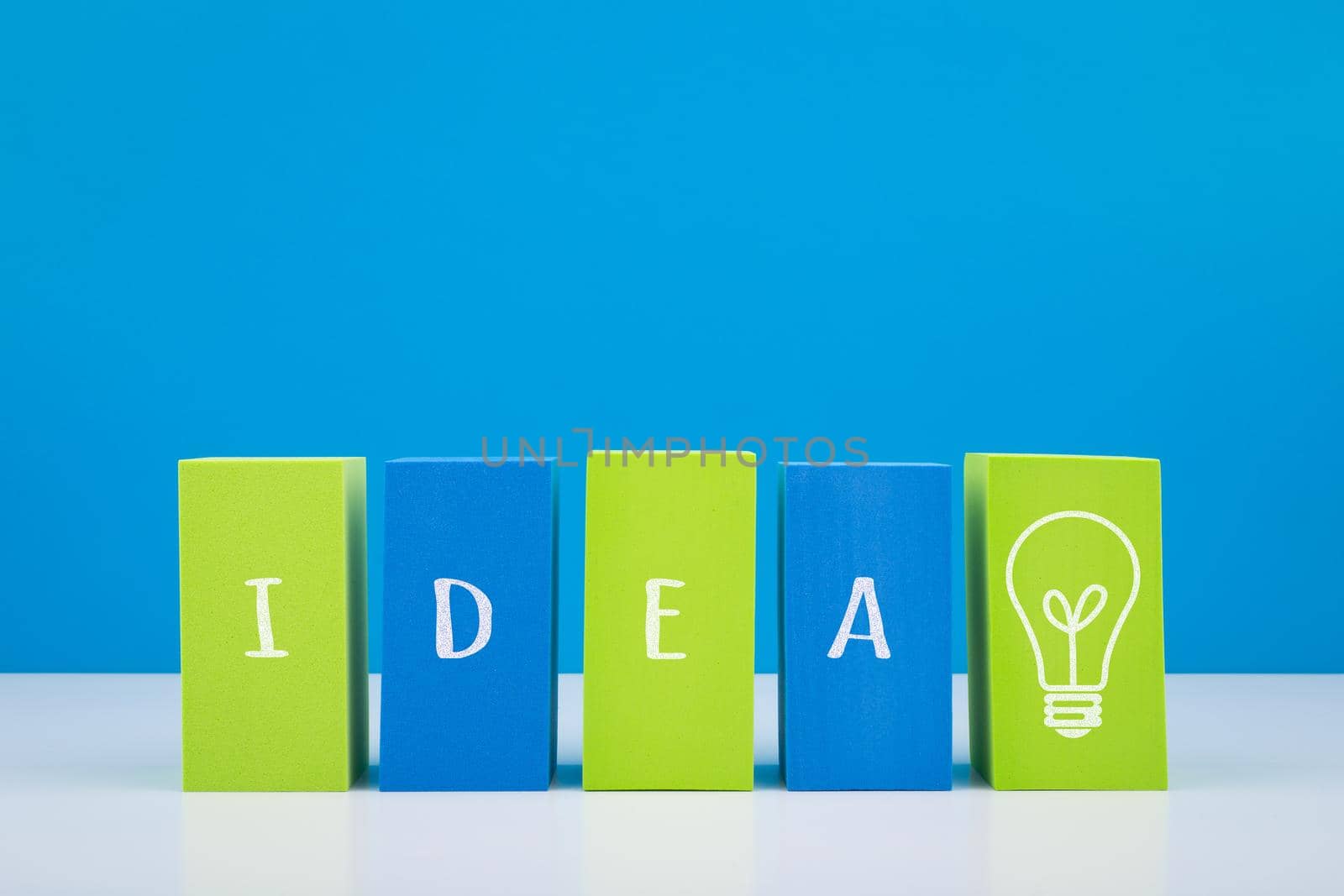Minimal colorful concept of brainstorming and new ideas. Blue and green blocks in a row with drawn word idea and light bulb on white table against bright blue background