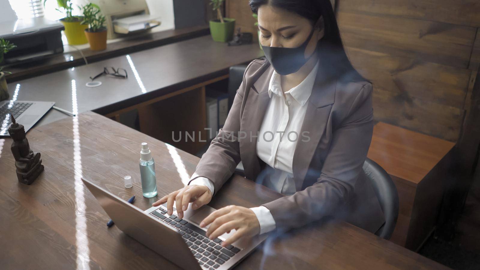 Asian Woman Working On Laptop During COVID 19 Pandemic, Businesswoman In Formalwear And Protective Mask Sitting At Office Table With Sanitizer Spray On It And Typing On Computer Keyboard, High Angle View