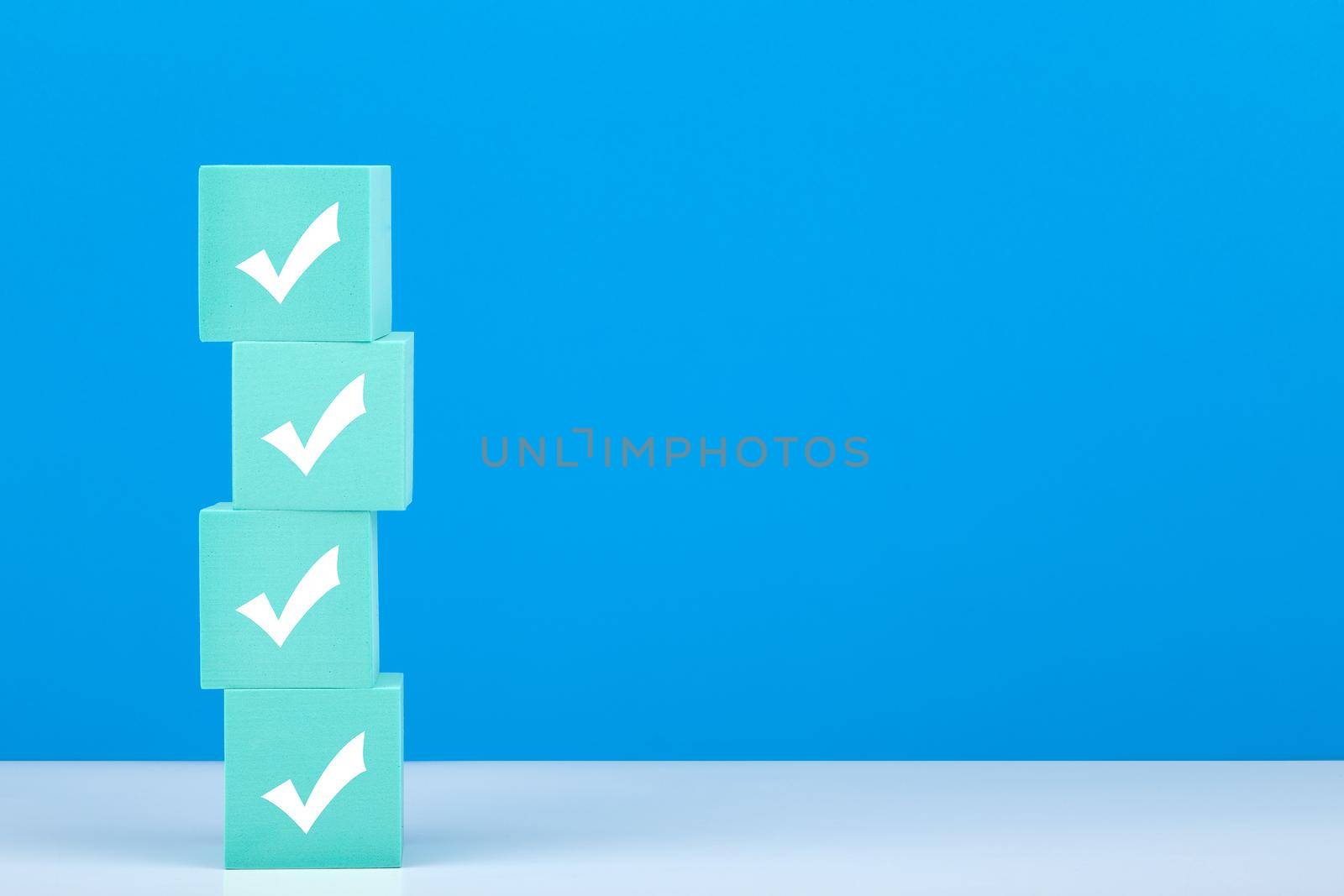 Four white checkmarks on stack of light blue toy blocks on white table against blue background with copy space. Concept of questionary, checklist, to do list, planning, business or verification