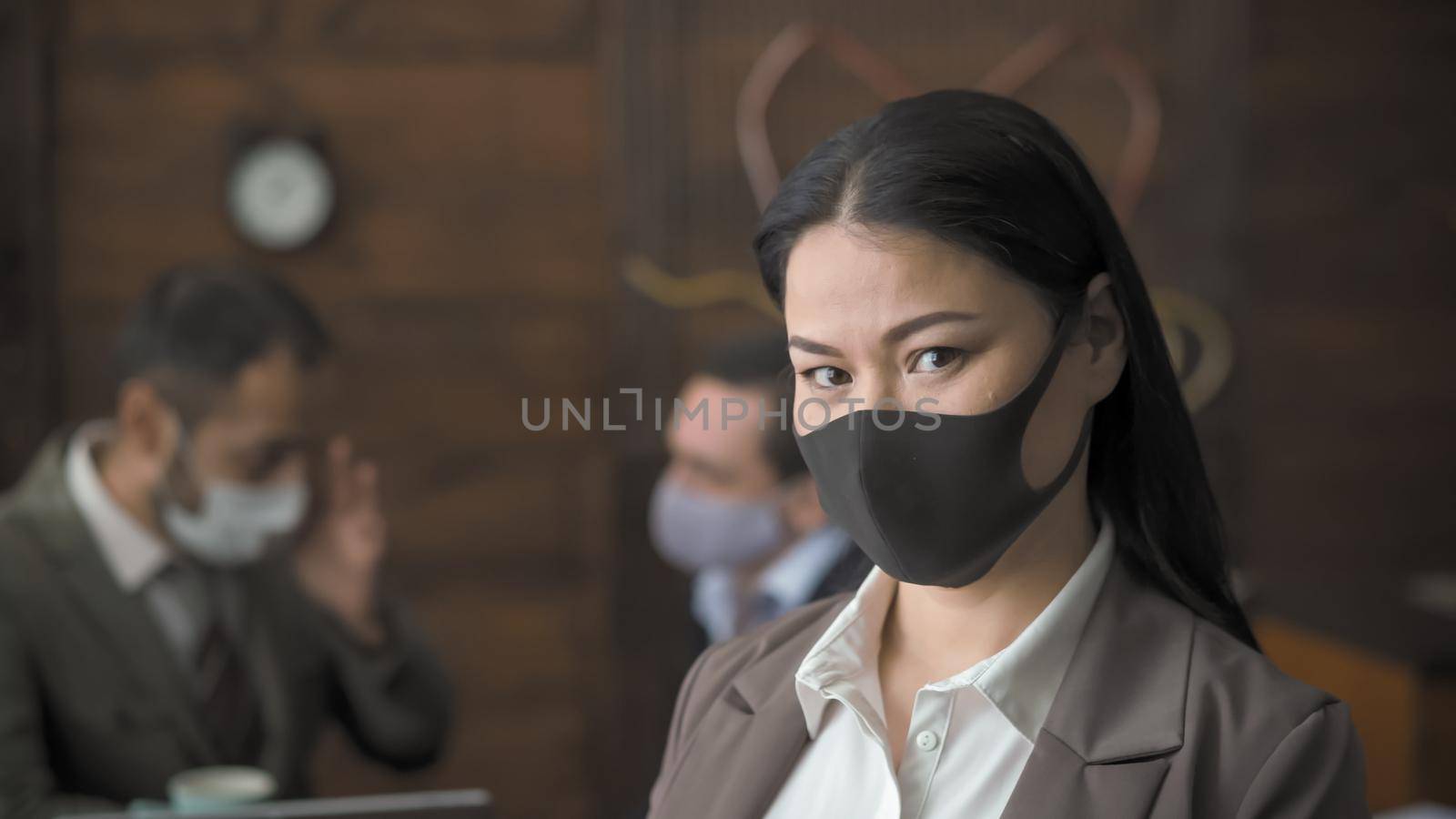 Brunette In Black Protective Mask Looks At Camera, Beautiful Asian Woman Is Standing In Office Against The Background Of Her Communicating Colleagues, Virus Outbreak Concept