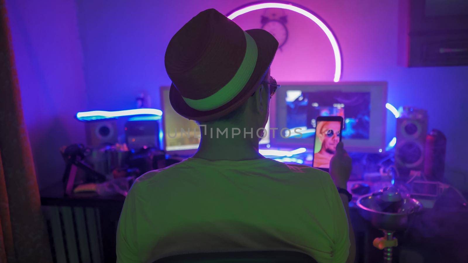 Man Smokes Hookah Making Selfie At Home During Quarantine, Rear View Of Man Smoking Hookah While Sitting At Work Place In Home Interior With Neon Lights, Self Isolation Concept