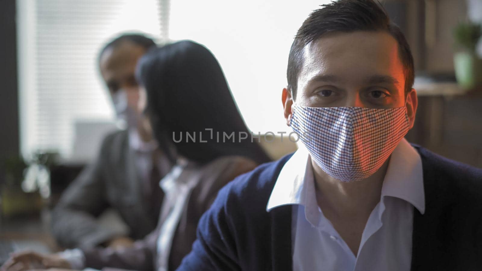 Business Partners Working During Quarantine, Focus On Caucasian Young Man In Protective Mask Looking At Camera In The Foreground, His Colleagues Communicate On Blurred Background.