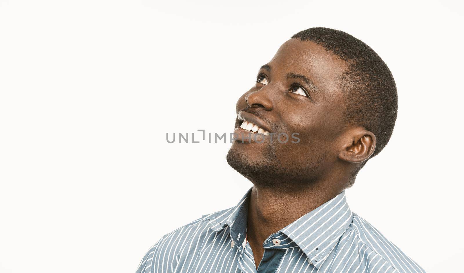 African american handsome man toothy smiles looking up and to the side, Dark-skinned young guy in blue striped shirt isolated on white background. Copy space located at left side. Toned image.