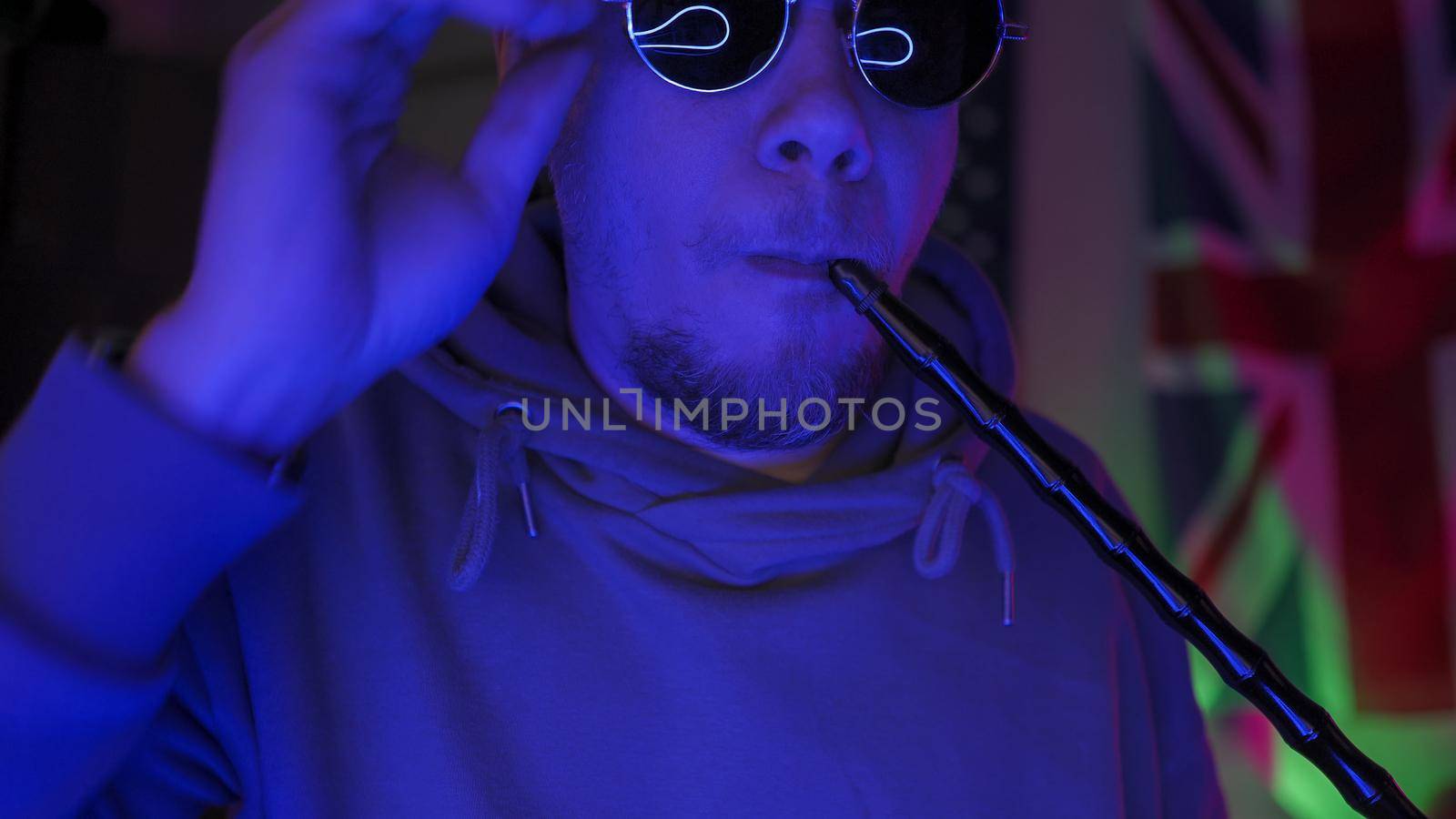 Hipsters Smoking Hookah Having Fun Self-Isolation Time At Home In Neon Backlight, British Man In Sweatshirt Corrects Sunglasses Looking At Camera Against The Background Of Great Britain Flag