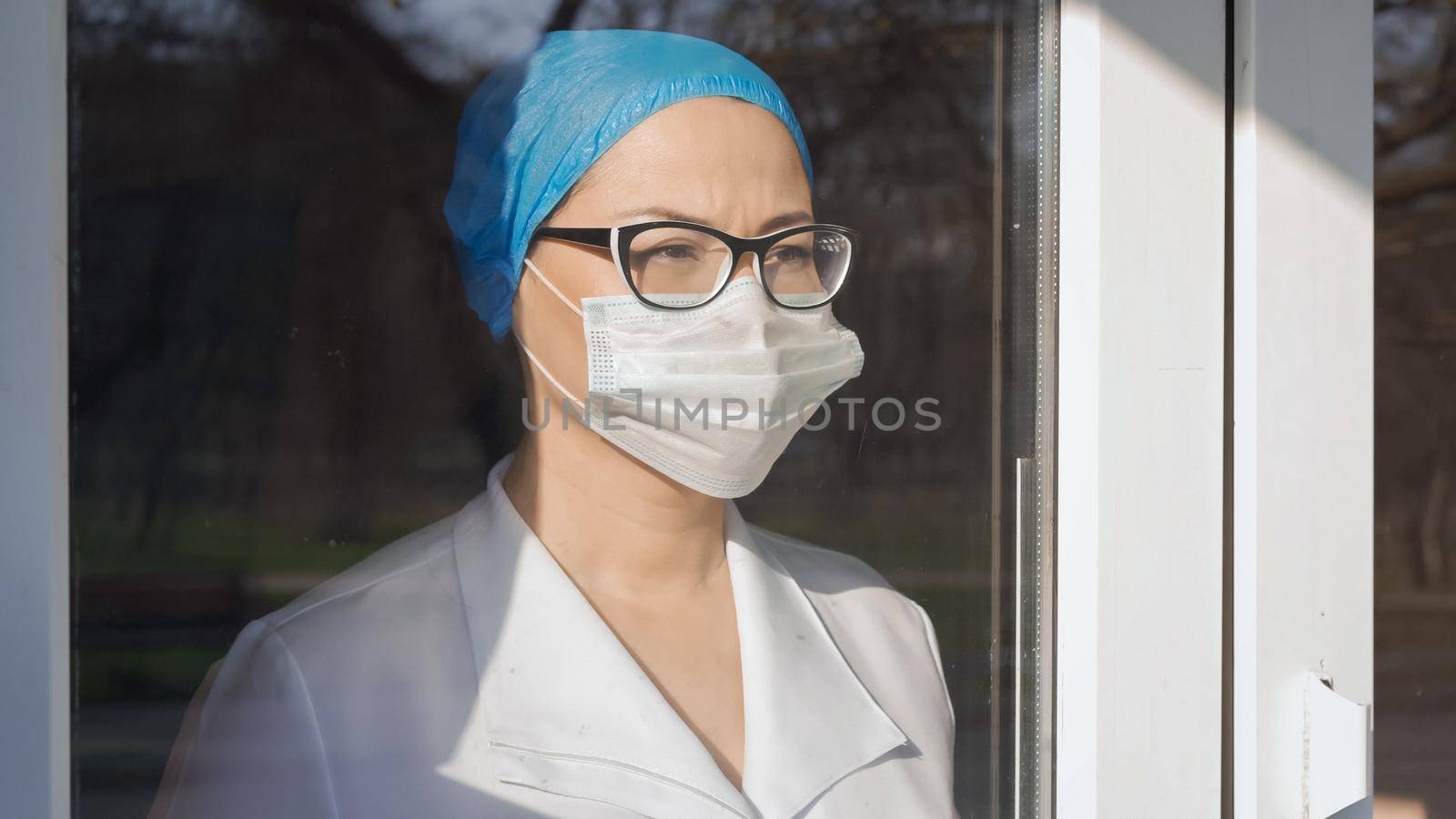 Female Medic Looks At Window Standing Behind Glass In Isolation On Sunny Day, Tired Woman In Protective Mask And Uniform Looks At Window Waiting For End Of Epidemic, Pandemic Concept