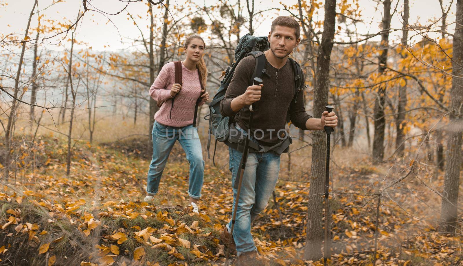 Traveling Couple Of Backpackers Hiking In Autumn Forest Outdoors, Caucasian Man Walks In Front Leaning On Hiking Poles And Looking Around, Blonde Woman Follows Him