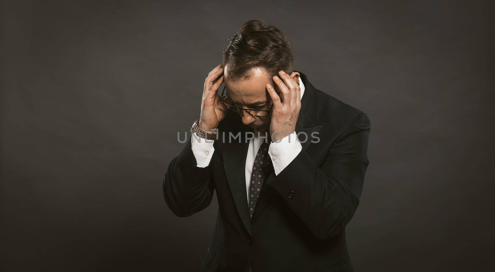 businessman in a depression holds his hands on his head. man in a suit with gray background clasped his head with his hands in a panic. Business problem concept by LipikStockMedia