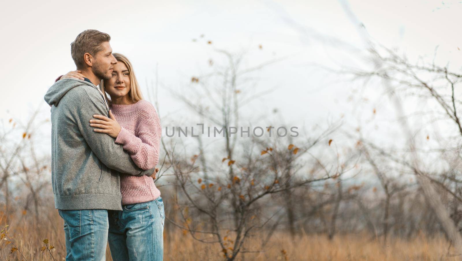 Loving couple resting while standing embracing on a background of autumn natural landscape. Young caucasian man and woman hugging on blurred young forest background. Copy space on the right side.