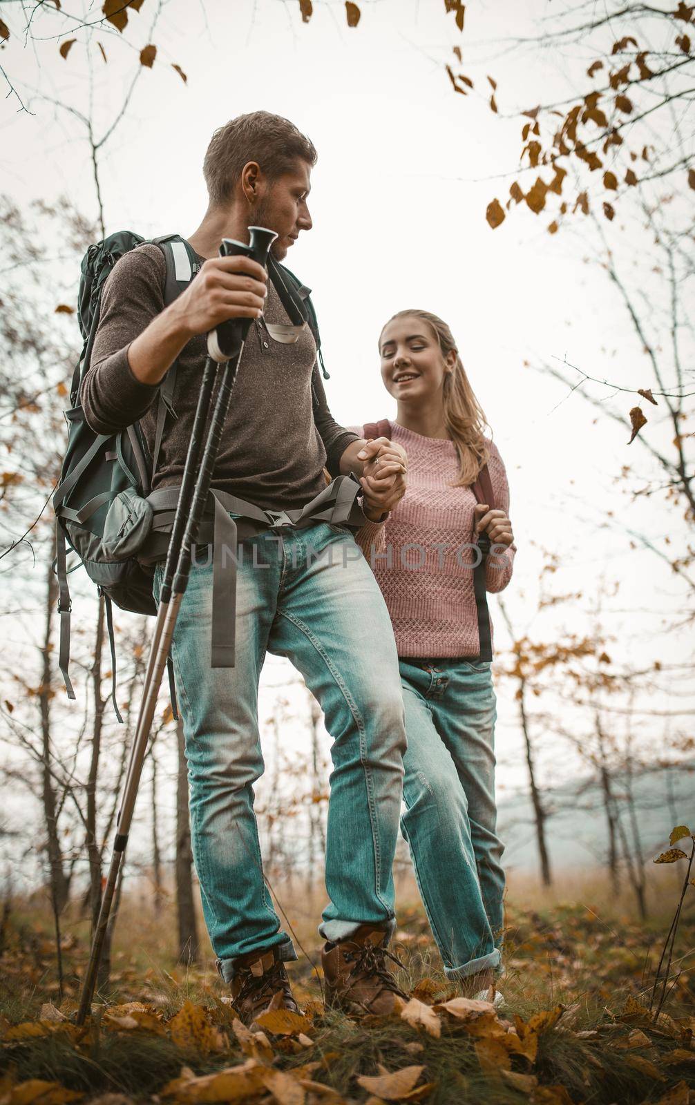 Tourists Backpackers Walking In Autumn Nature On Fallen Leaves And Green Grass, Focus On Man Holding His Woman's Hand And Hiking Poles In Other Hand, Walk With Backpacks Concept, Shot From Below