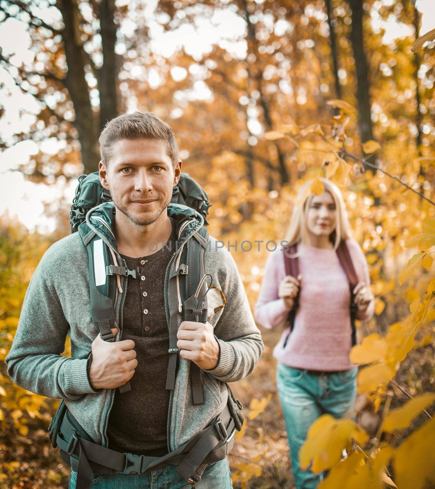 Charming Couple Of Travelers With Backpacks Walking Through The Autumn Forest, Selective Focus On Caucasian Man Standing In The Foreground, Beautiful Blonde Follows Him