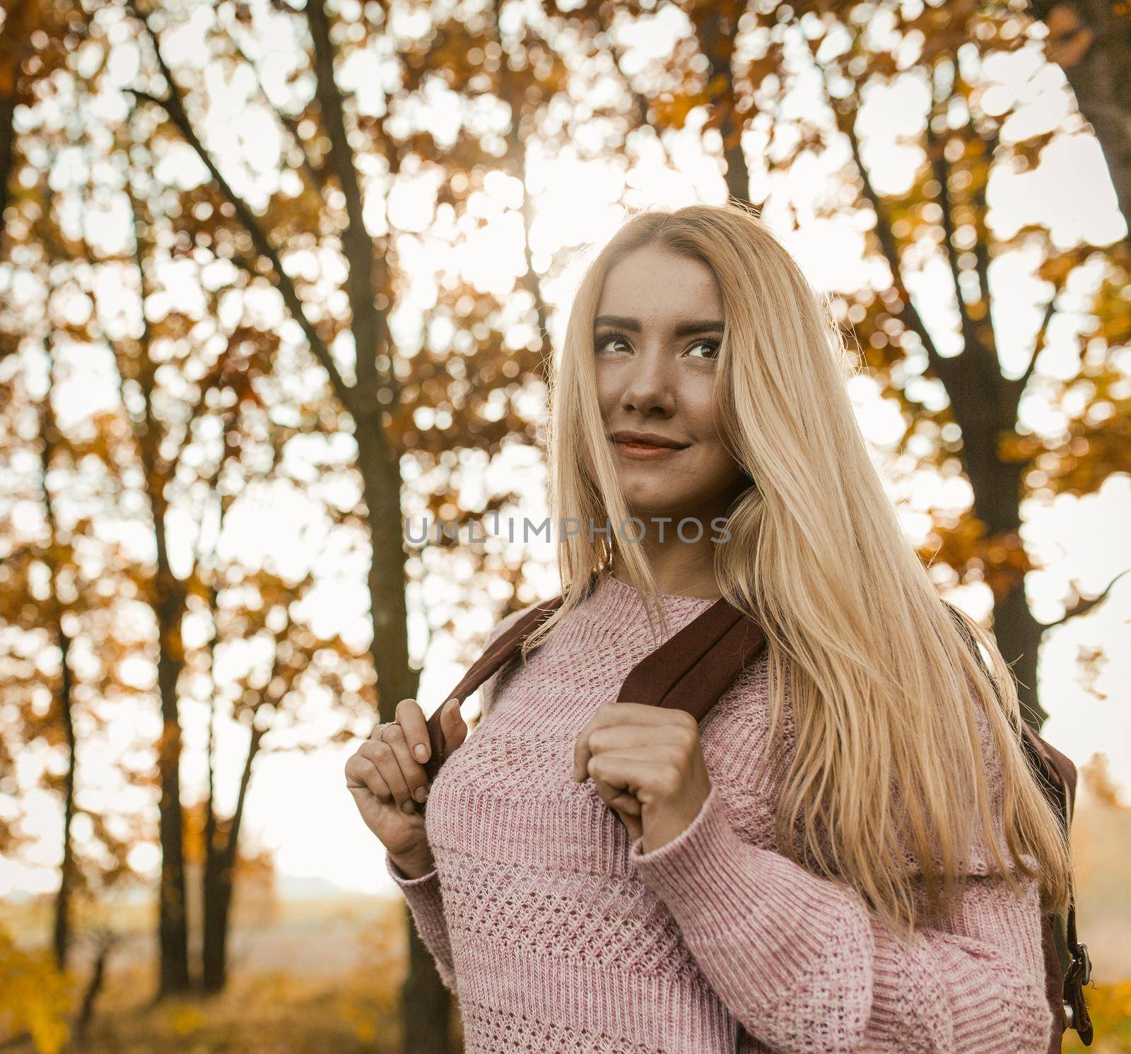 Charming Blonde Walking In Autumn Forest, Portrait Of Cheerful Young Woman With Long Hair Holding Backpack With Her Hands And Looking Up Against Background Of Autumn Forest, Healthy Lifestyle Concept