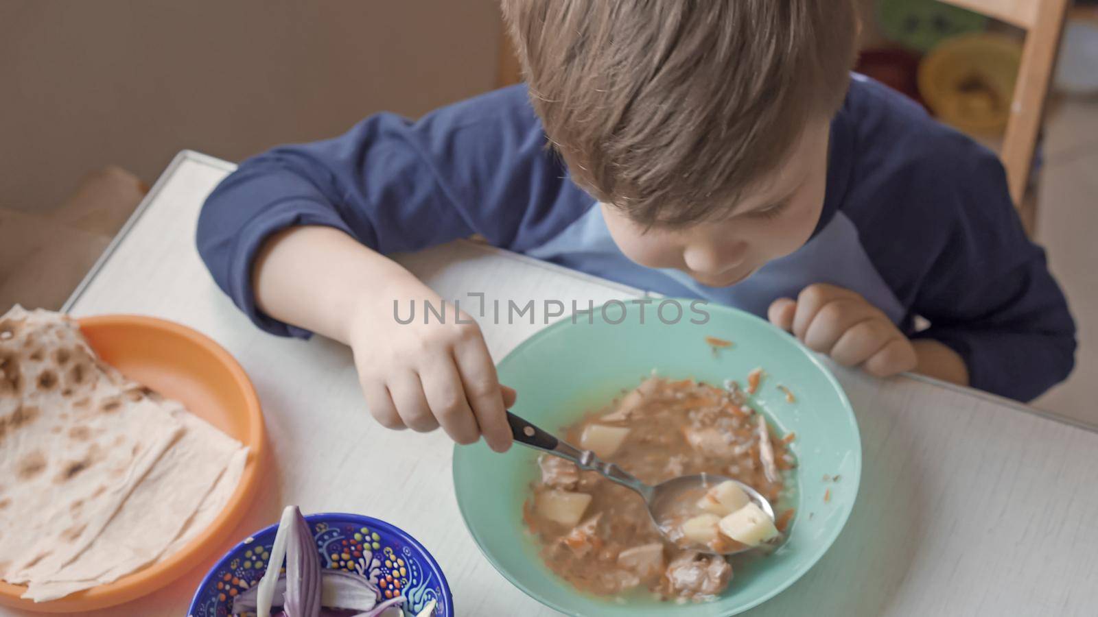 Hungry Boy Sits At Table Eating Soup By Himself by LipikStockMedia