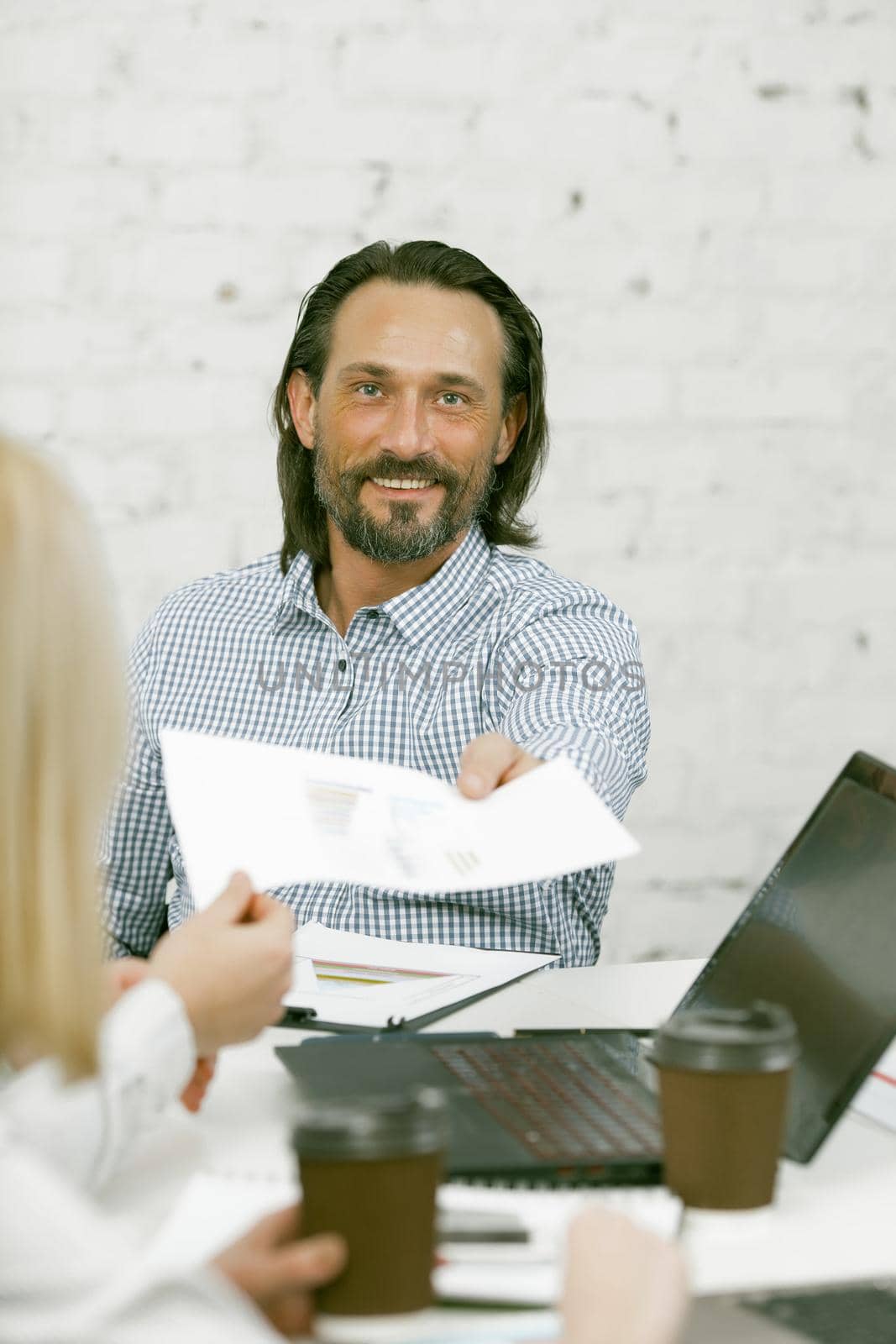 Smiling Businessman passes the paper form or sheet of paper to his colleague. Bearded caucasian man looks at camera sitting at meeting table in light modern office. Toned image.