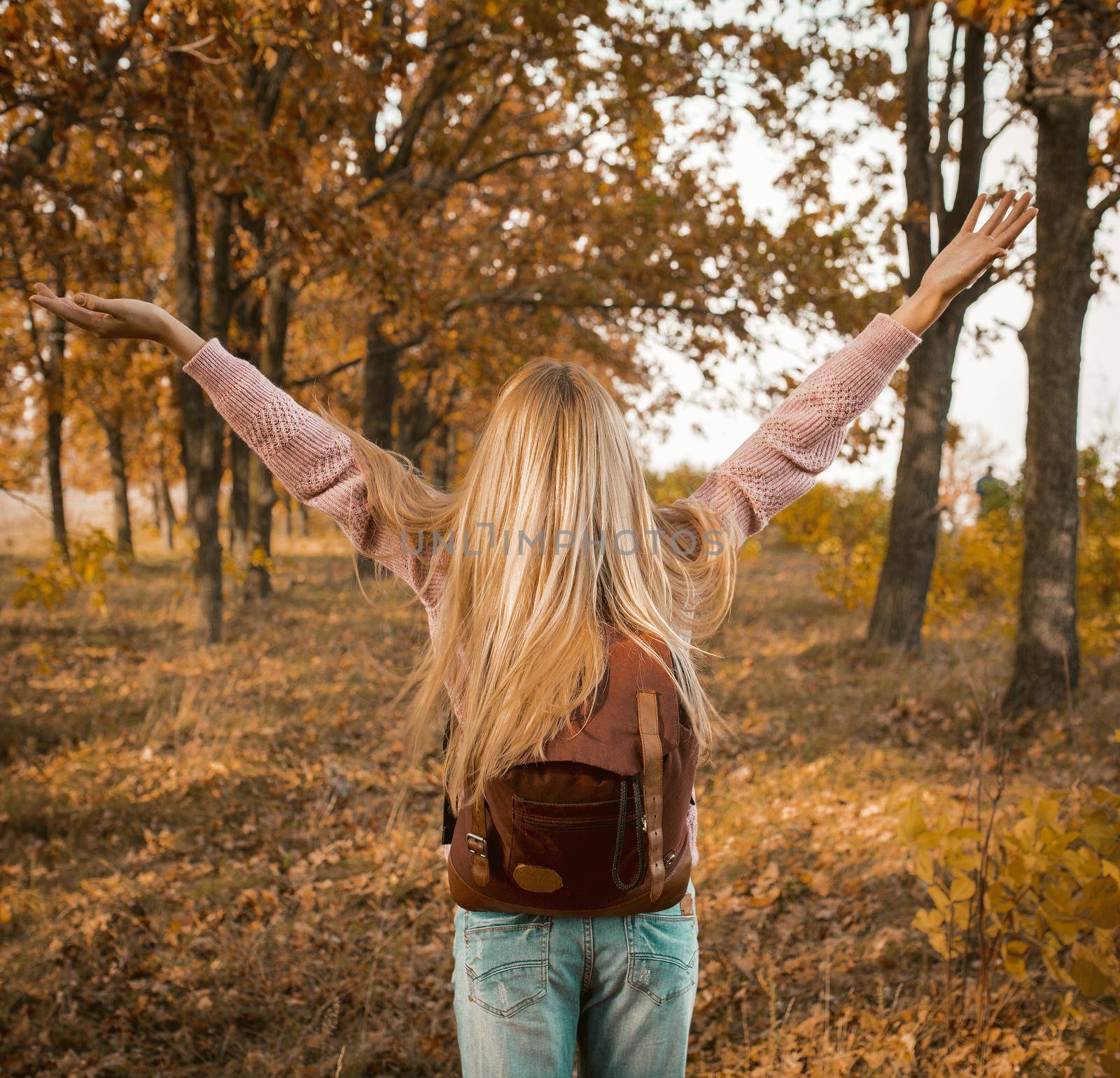 Happy Woman With A Backpack Stands Against The Backdrop Of The Autumn Forest, Raising Her Arms To Sides, Rear View Of Young Caucasian Blonde With Long Hair Wearing Jeans Standing In Nature In Sunlight
