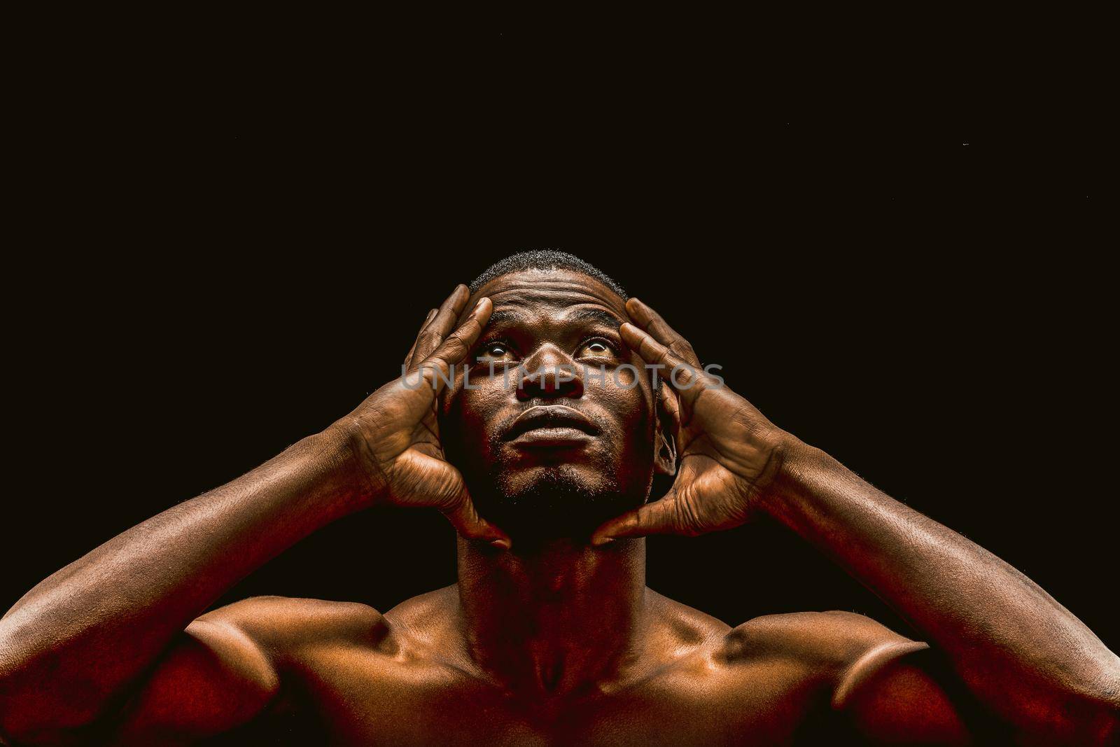 Handsome African american man looking up touching hands of head, portrait of nude dark-skinned athlete isolated on black background. Copy Space is at the top of Image. Toned image by LipikStockMedia