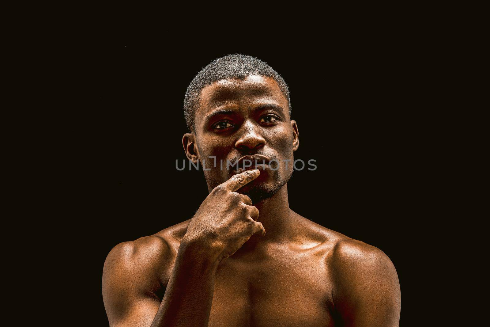 Naked African man thoughtfully looks at the camera while touching the fingers of his lips, sexy muscular young guy posing in the studio, cut out on a black background. Toned image by LipikStockMedia