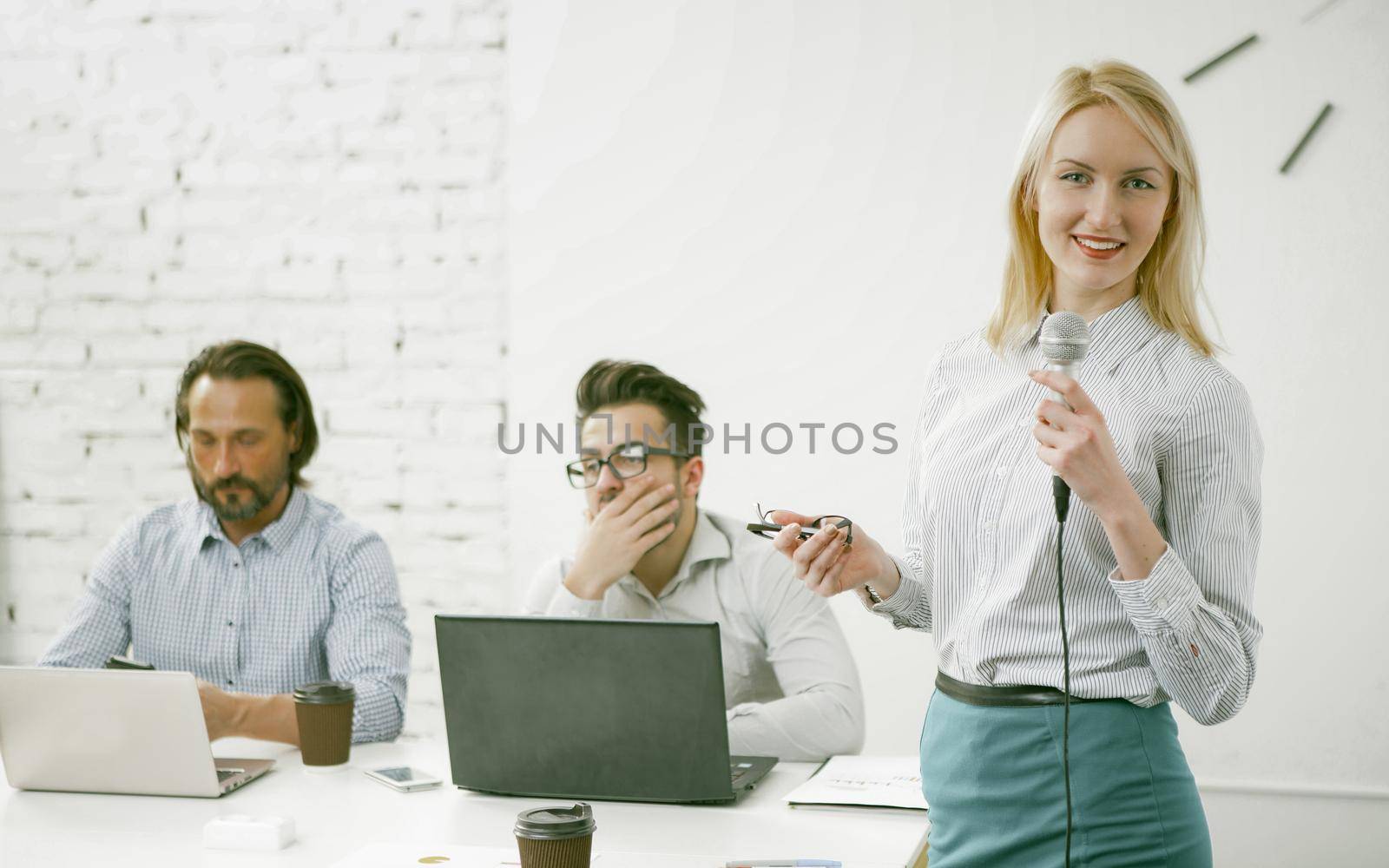 Smiling business woman speaks into microphone, Charming blonde shares ideas about company development while standing in foreground against background of colleagues sitting at office desk. Toned image.