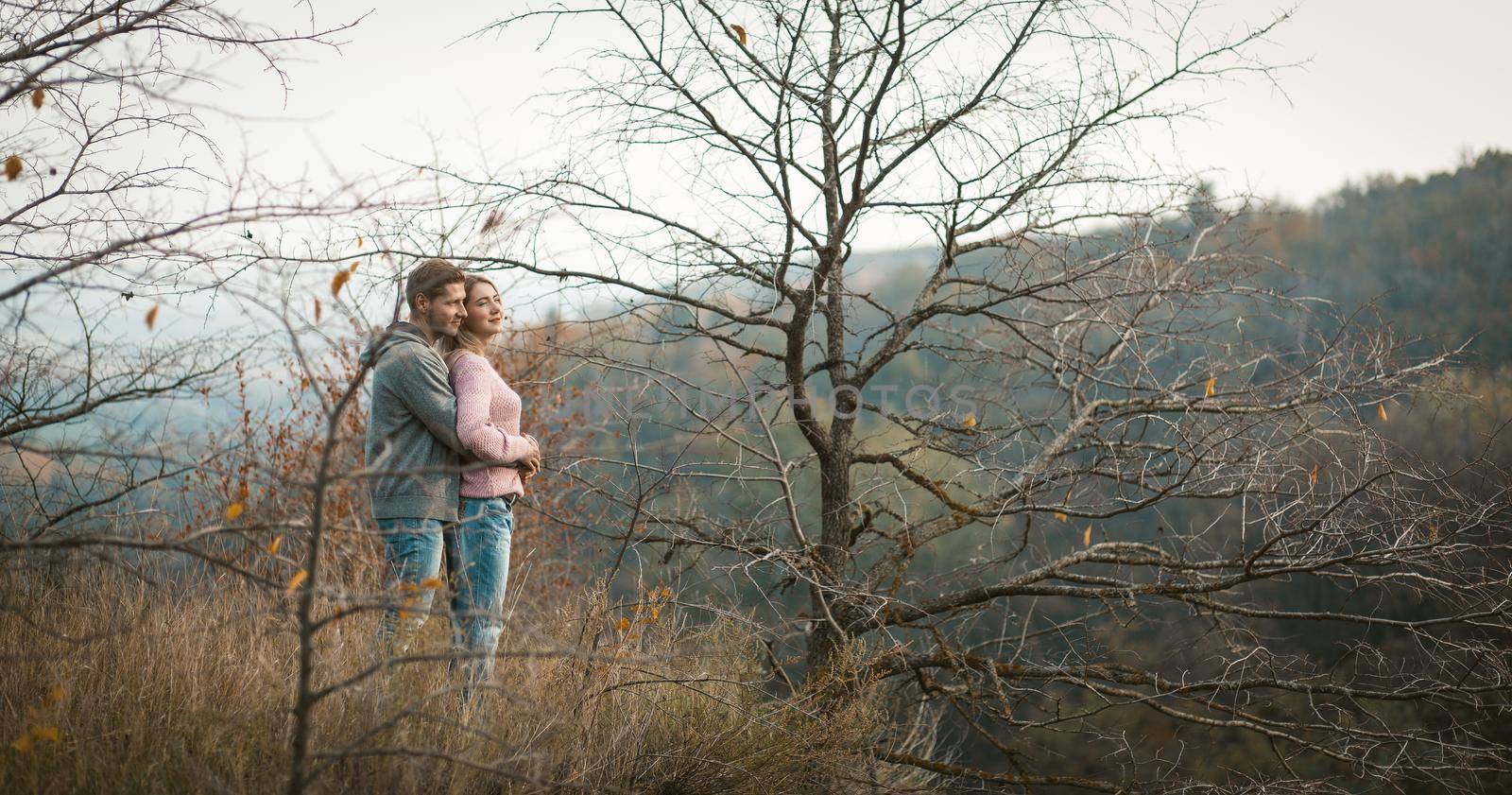 Hugging couple in love stands on a hillside looking down, young man and a woman admire the autumn forest growing on the slopes below, travel on nature concept