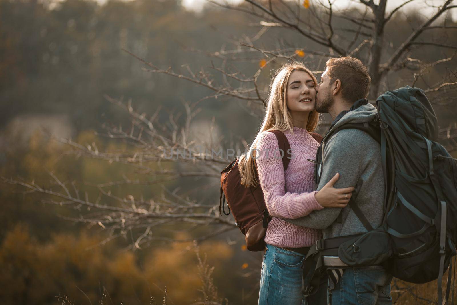 Men and women with backpacks stand gently embracing against the backdrop of autumn nature on the edge of a cliff. Young man kisses his beautiful blonde wife outdoors.