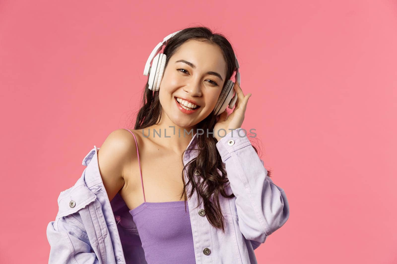 Close-up portrait of charismatic good-looking korean girl enjoying listening music, wear headphones smiling and dancing, found great playlist for walking on fine spring day, pink background.