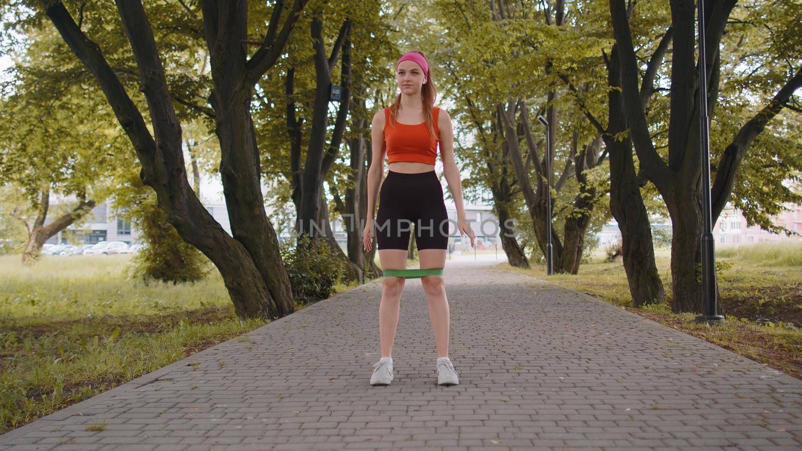 Athletic fit slim sport girl doing squats using fitness elastic rubber band as part of her fitness workout outside in park at morning. Young woman enjoying healthy lifestyle. Active sportswoman jogger