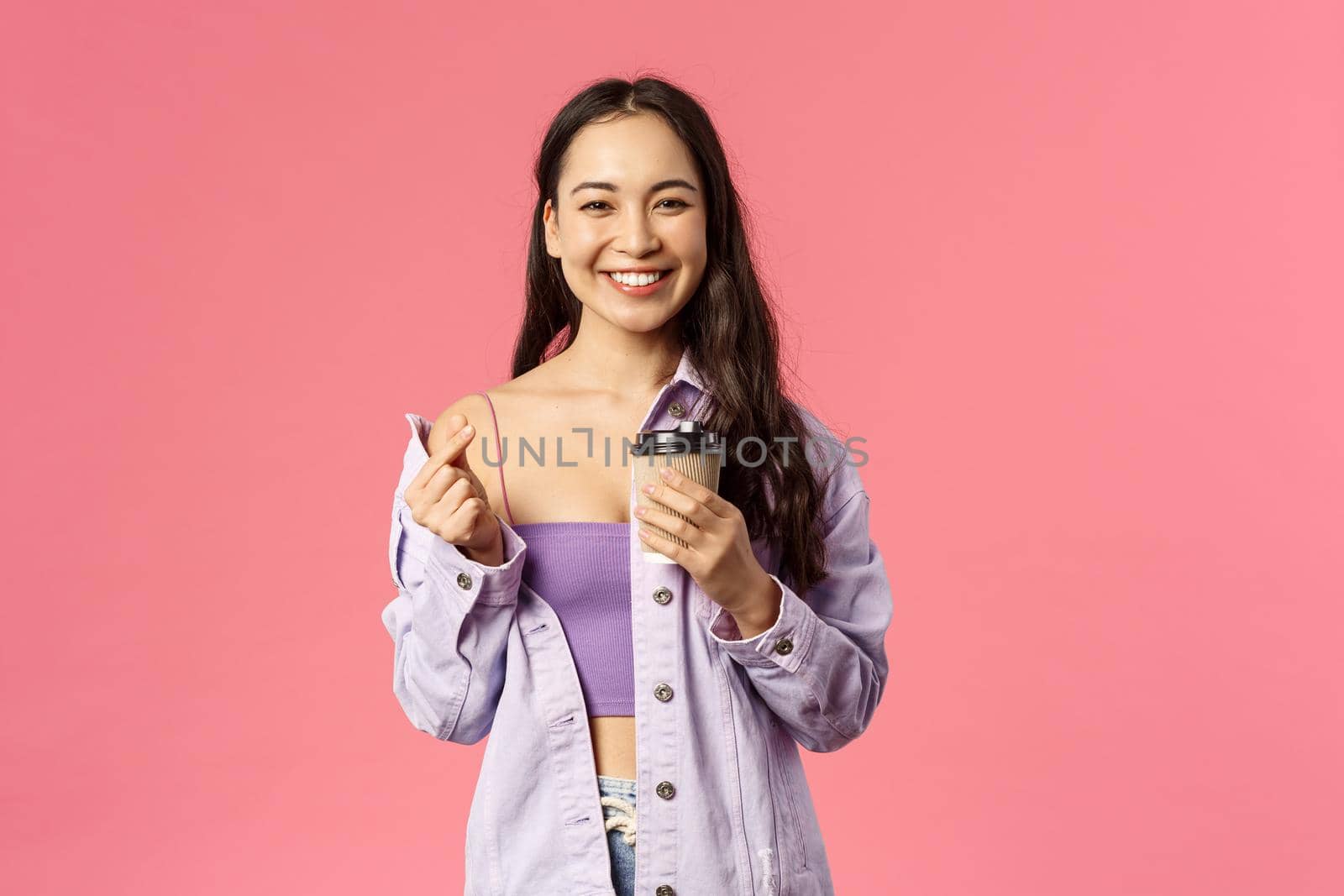 Happy stylish young modern female student walking from her favorite cafe, holding take-away coffee cup and smiling, drinking beverage enjoy sunny spring day, show love heart sign, pink background.