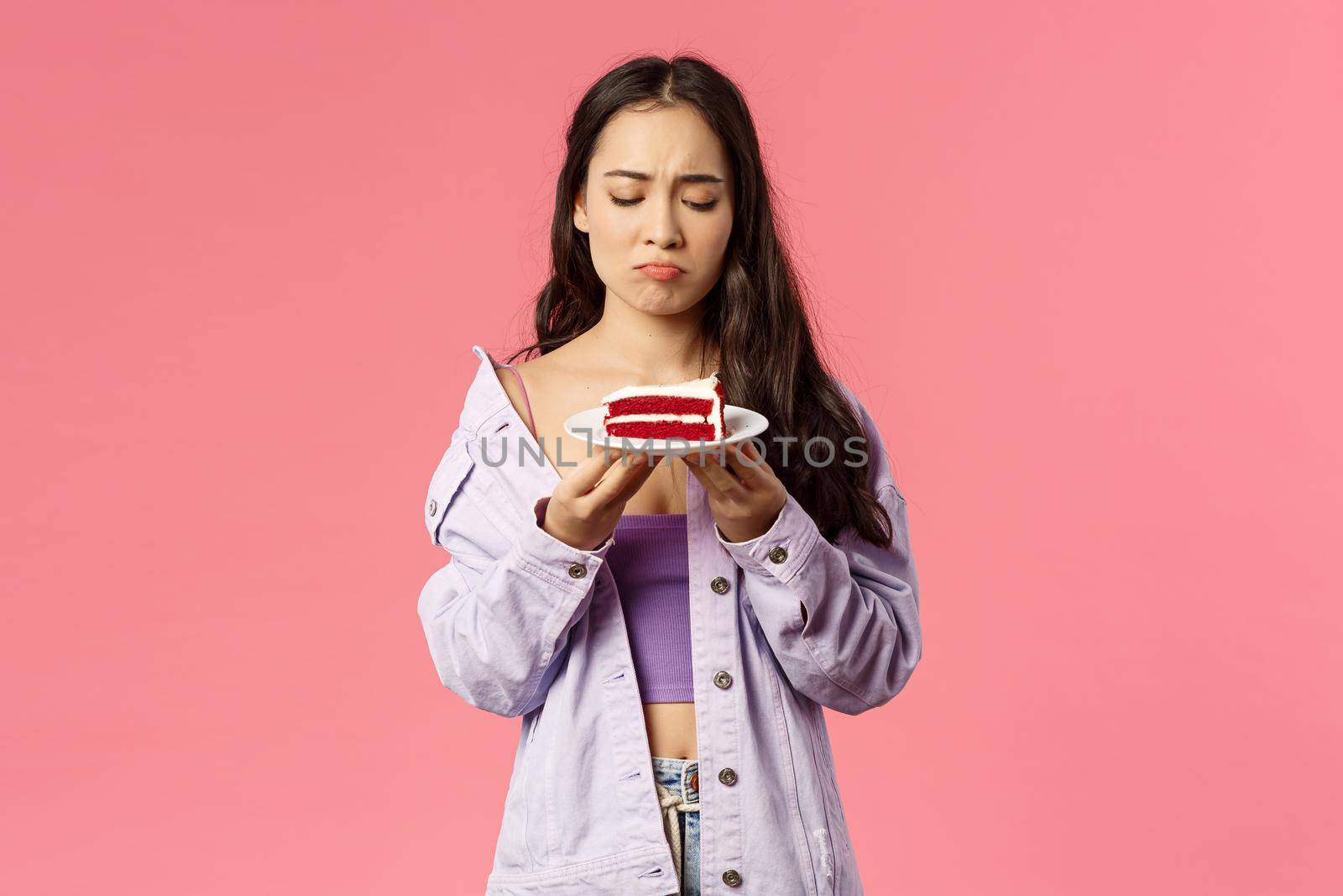 Portrait of gloomy upset asian female sighing, look uneasy and distressed at delicious piece cake but cant eat it, express regret and disappointment, standing pink background distressed.