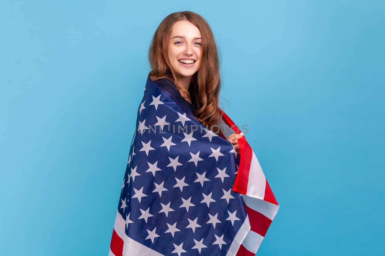 Portrait of satisfied woman wearing striped casual style sweater, standing wrapped in american flag, celebrating National independence day - 4th july. Indoor studio shot isolated on blue background.