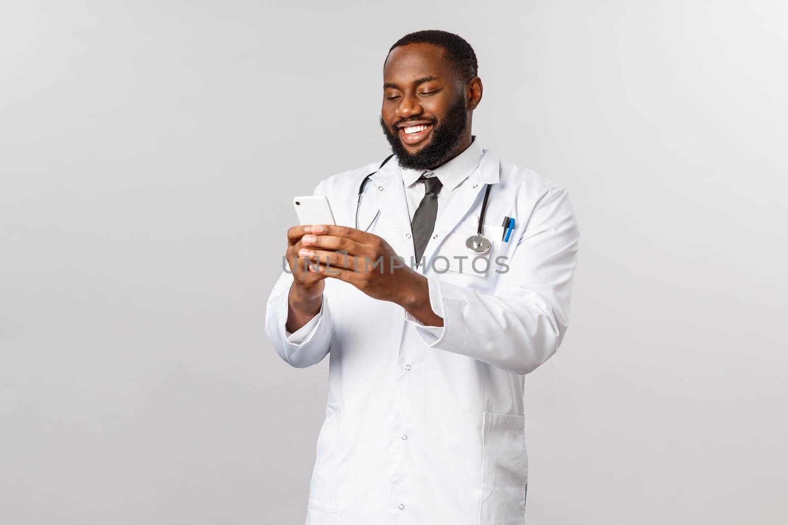 Covid19, pandemic and online medicine concept. Happy satisfied african-american doctor working hard in hospital during outbreak coronavirus, look at mobile phone, videcalling family with smartphone.