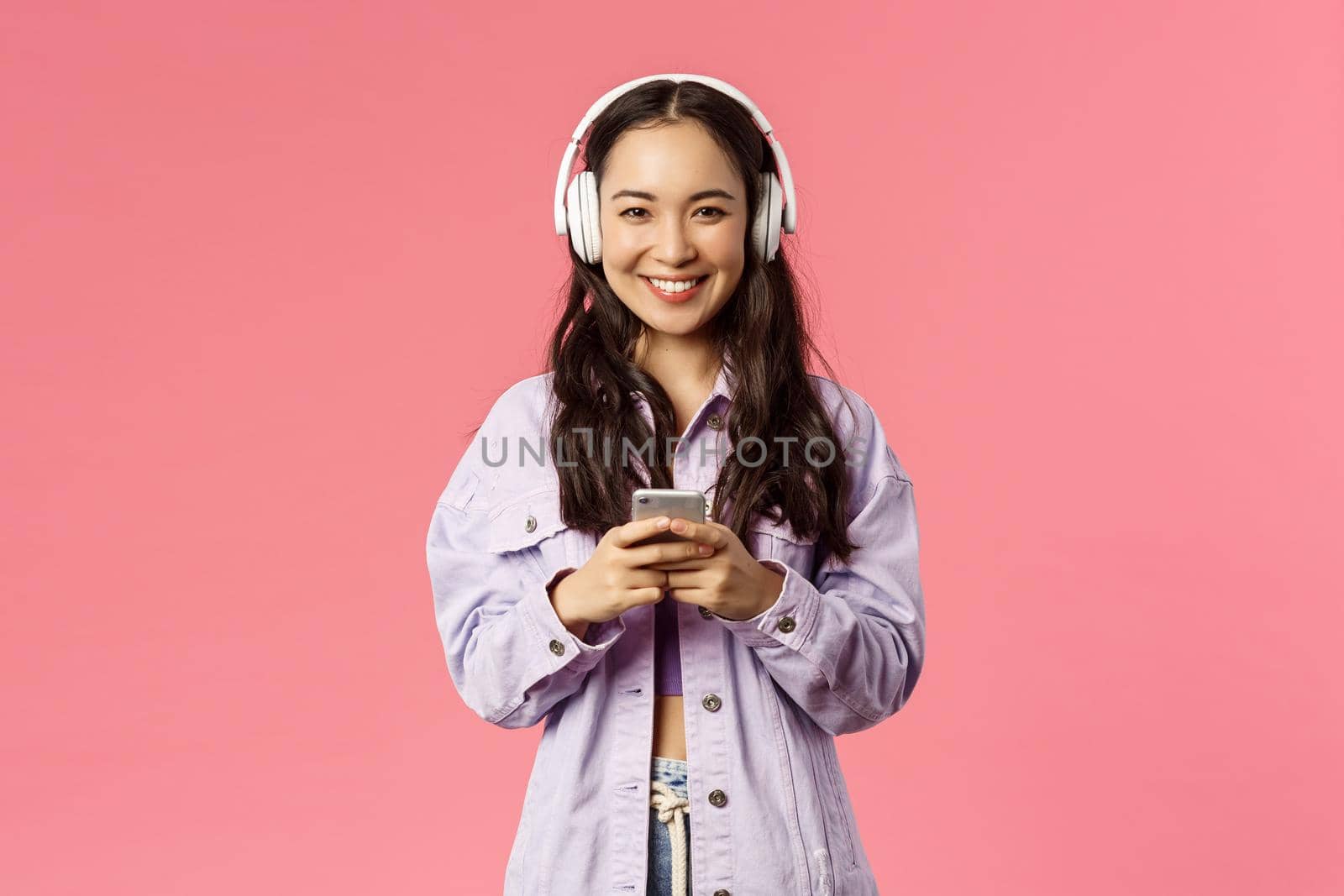 Portrait of attractive modern and stylish brunette korean, asian girl in headphones using mobile phone, listening music, made new playlist for going out, standing pink background.
