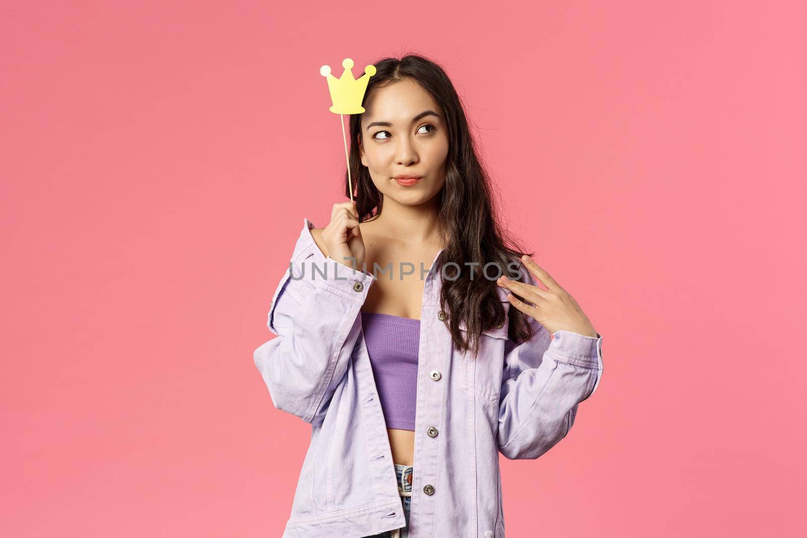 Holidays, lifestyle and people concept. Portrait of sassy, confident attractive asian girl looking thoughtful away, holding a crown stick over head, feel powerful like queen, pink background.