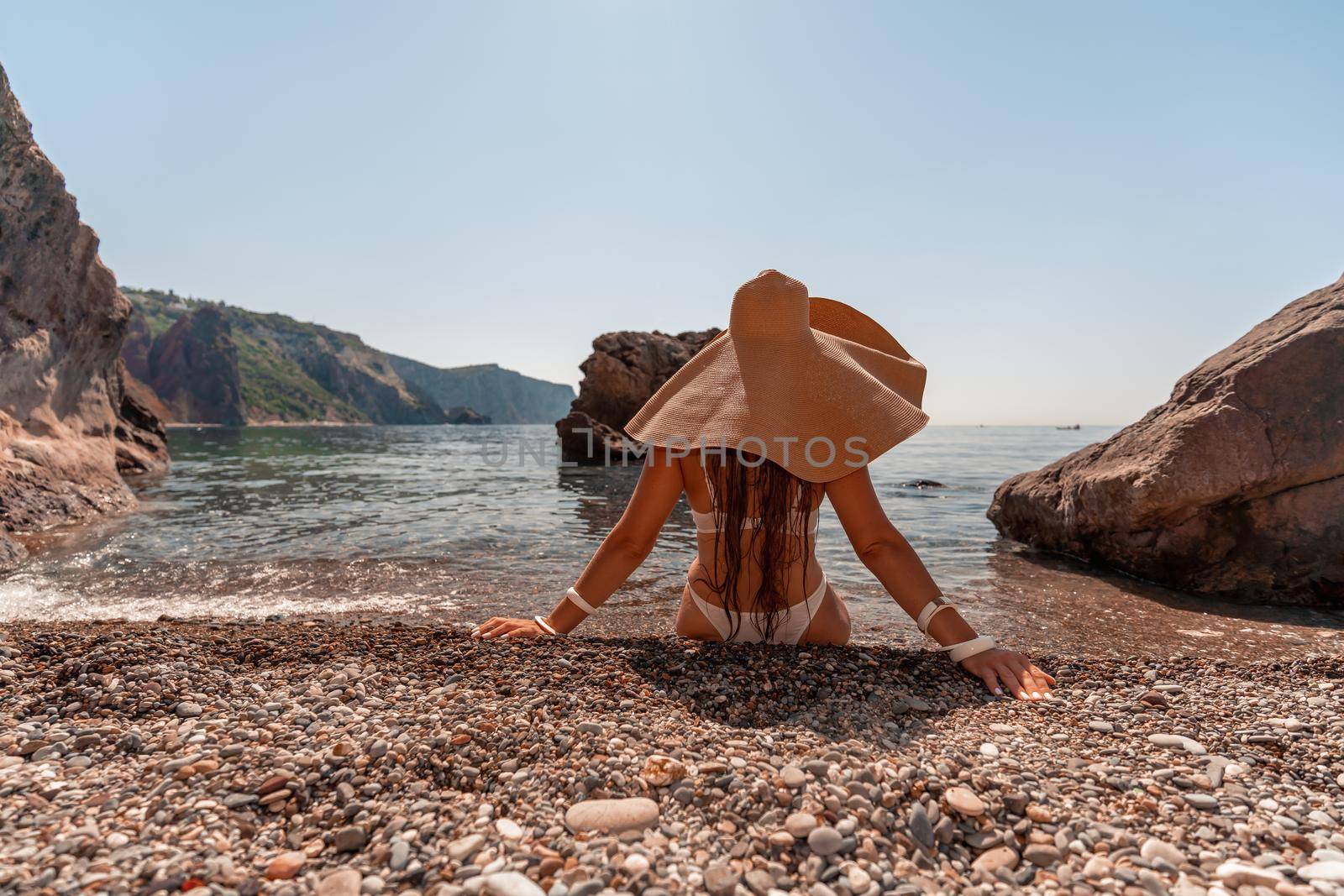 Tanned middle-aged woman with long hair and a white bathing suit. He sits on the seashore in a large sun hat with his back and looks at the sea. by Matiunina