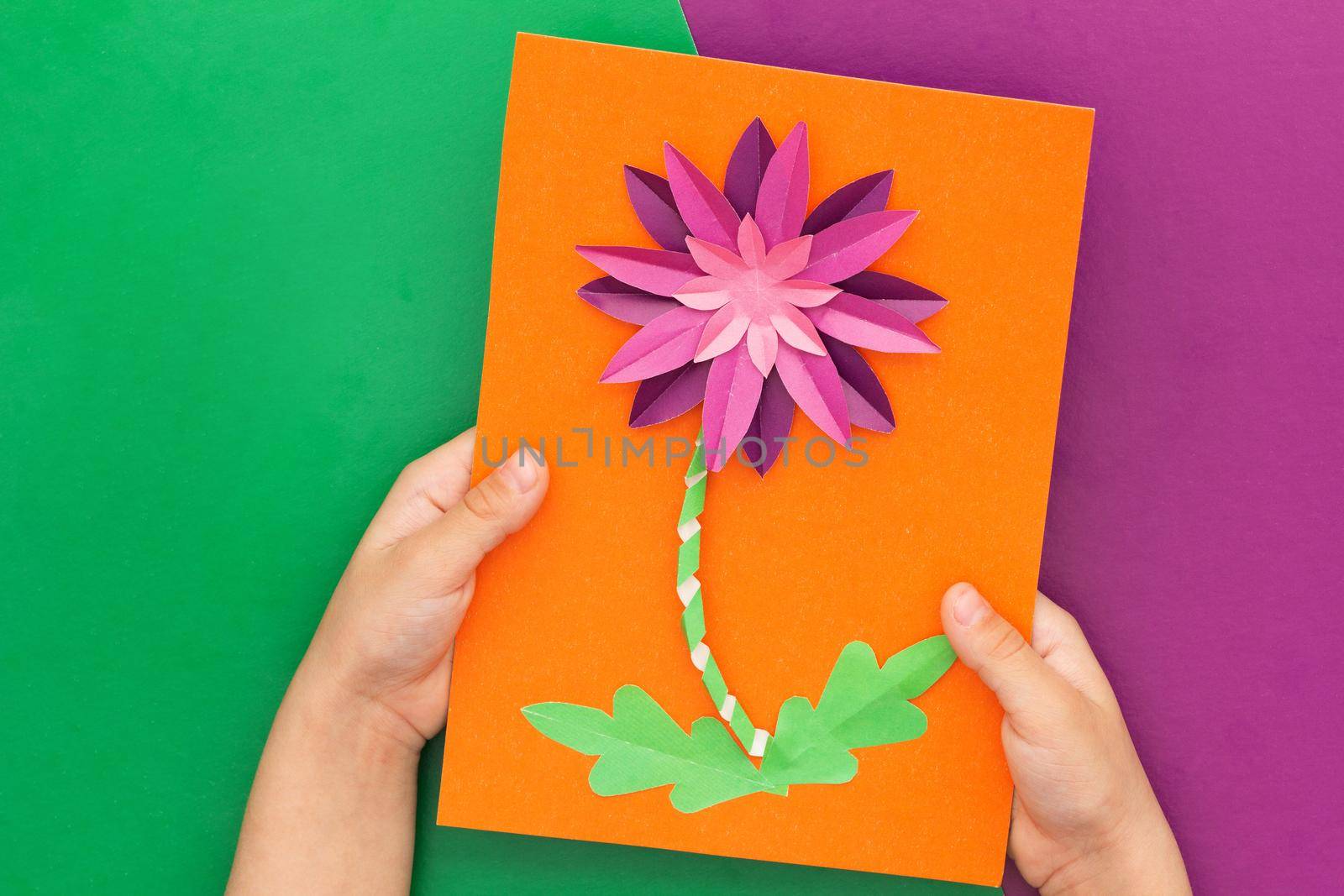 Top view of child hands holding papercraft violet crysanthemum flower made by child on orange worksheet on violet and green background. Present for mother s day