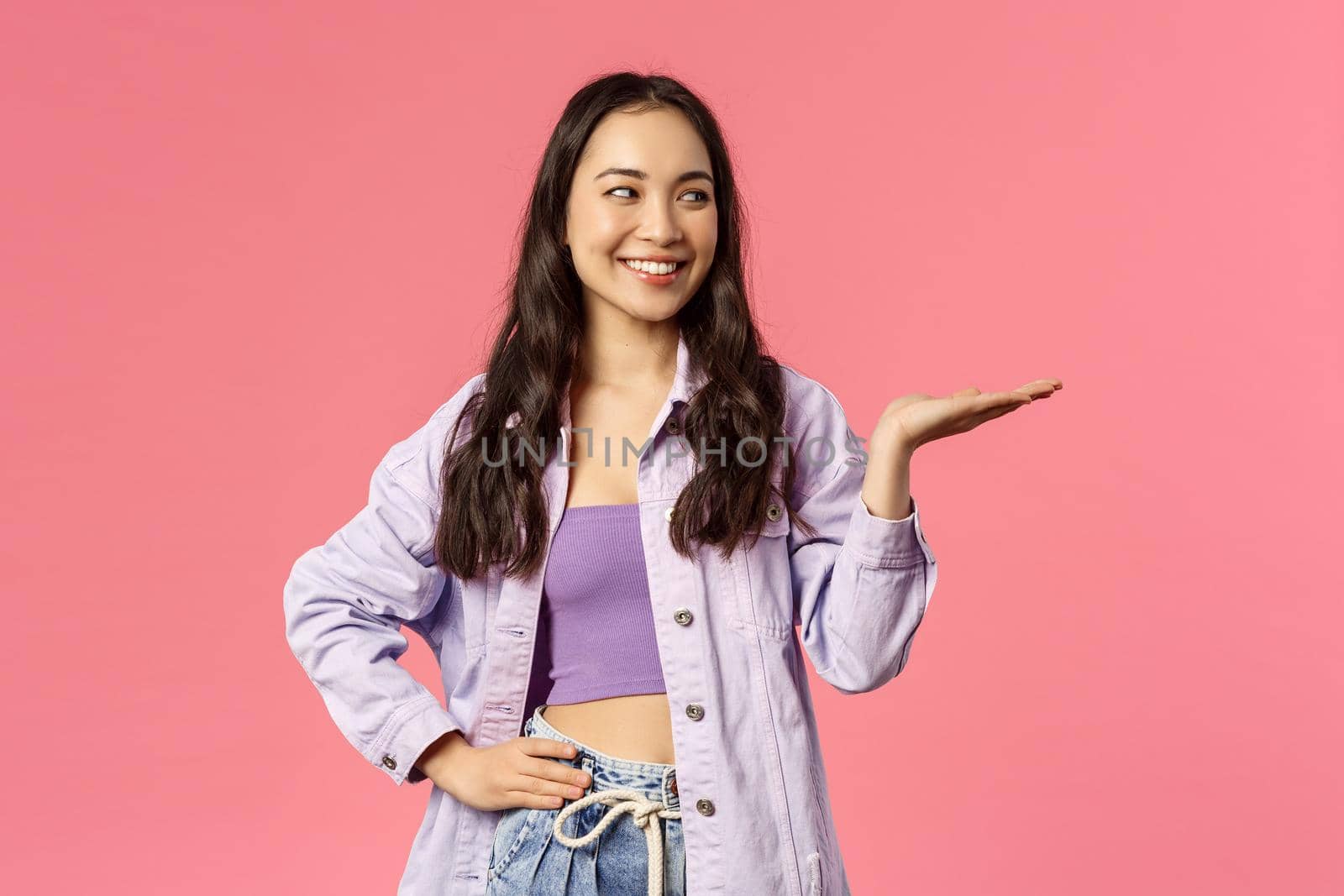 Girl presents you something worth your attention. Portrait of cheerful attractive asian female introduce product, pointing with palm and looking right, smiling pleased, pink background.