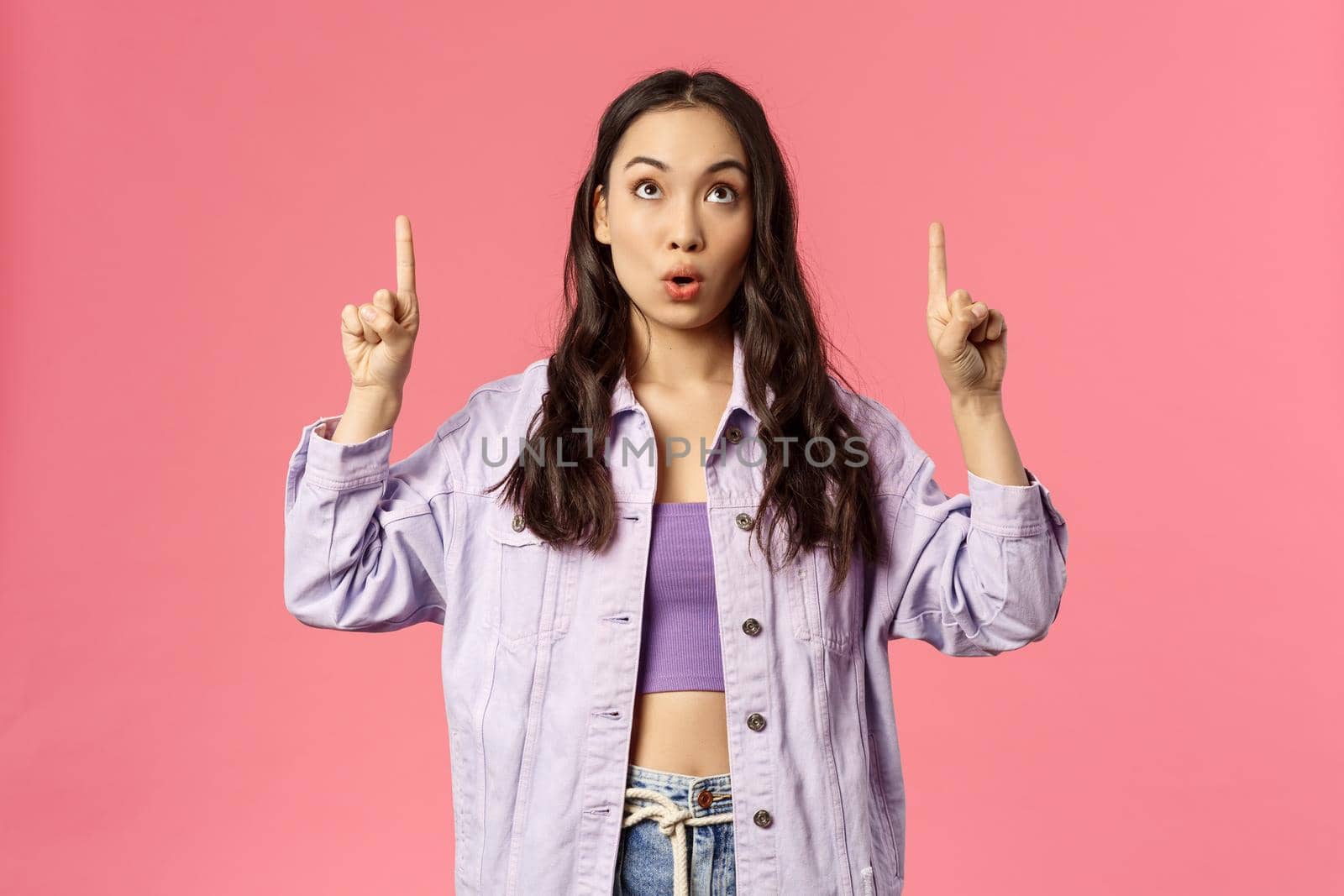 Portrait of curious and surprised young interested woman, mixed-race girl peeking and pointing fingers up, say wow, reading sign, visit interesting lecture or event, standing pink background by Benzoix